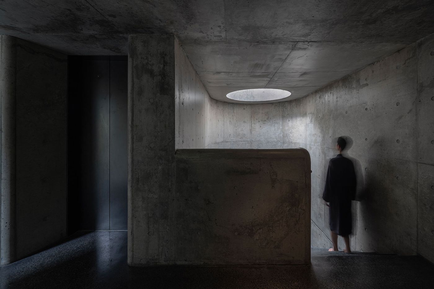 A figure in a dark robe stands in a minimalist concrete space, illuminated by a circular skylight above. A figure in a dark robe stands in a minimalist concrete space, illuminated by a circular skylight above.