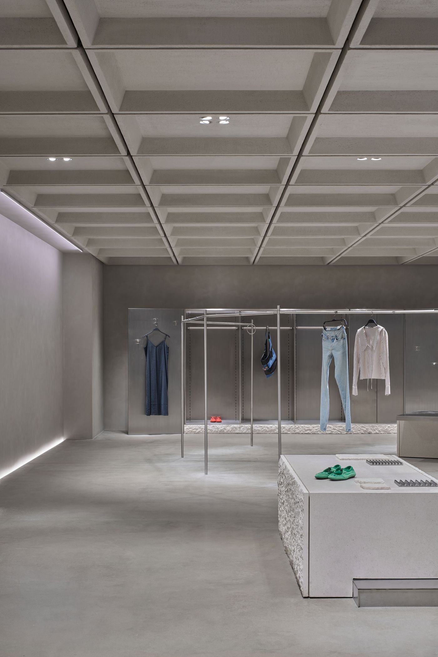 Minimalistic clothing store interior featuring hanging garments, modern concrete textures, and stylish green shoes on a display.