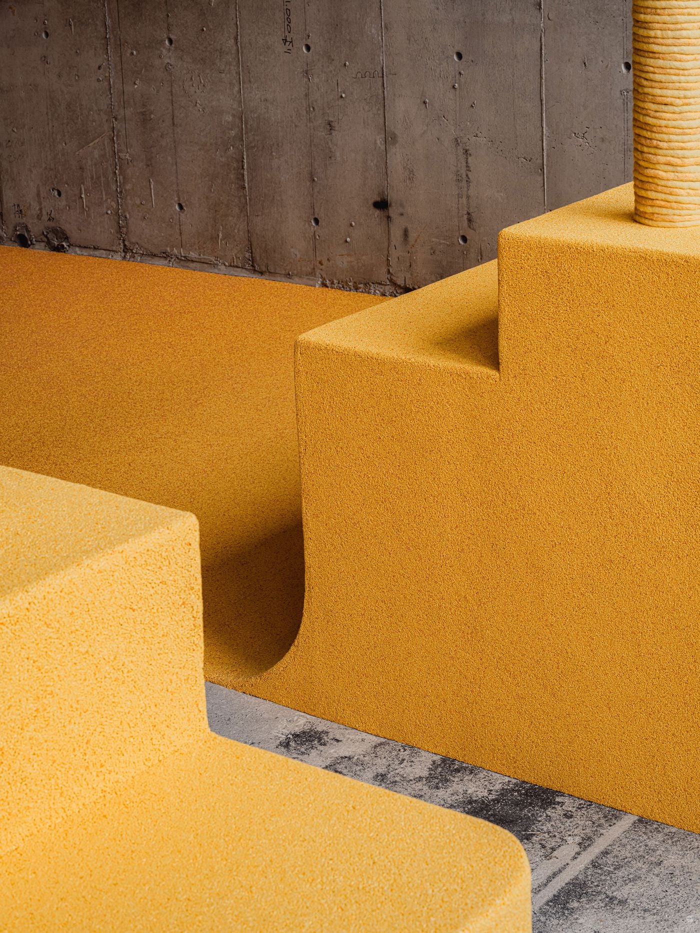 Canary-yellow microcement folds from floor into stepped display plinths and bench-like forms, their rounded transitions contrasting with the rough concrete wall behind. A rope-wrapped column rises from the composition, adding another tactile layer to I IN’s interplay of industrial structure, saturated colour and softer geometry.