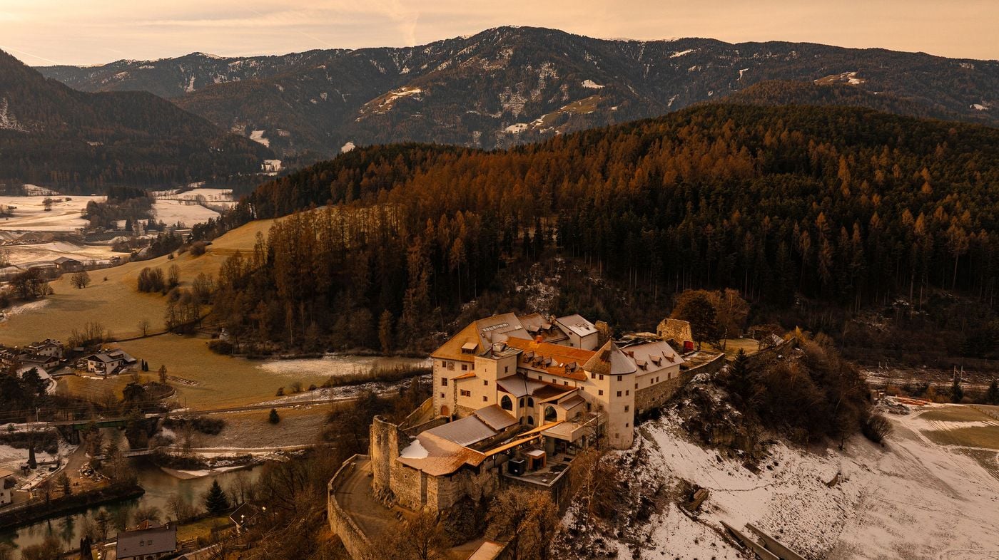 An aerial view at dusk shows Castel Badia's full complex on its rocky spur: rendered wings, a conical-roofed round tower, curtain walls and a fragment of medieval ruin. Russet larch forest rises behind, while a river, a railway line and frosted fields trace the valley floor below. An aerial view at dusk shows Castel Badia's full complex on its rocky spur: rendered wings, a conical-roofed round tower, curtain walls and a fragment of medieval ruin. Russet larch forest rises behind, while a river, a railway line and frosted fields trace the valley floor below.