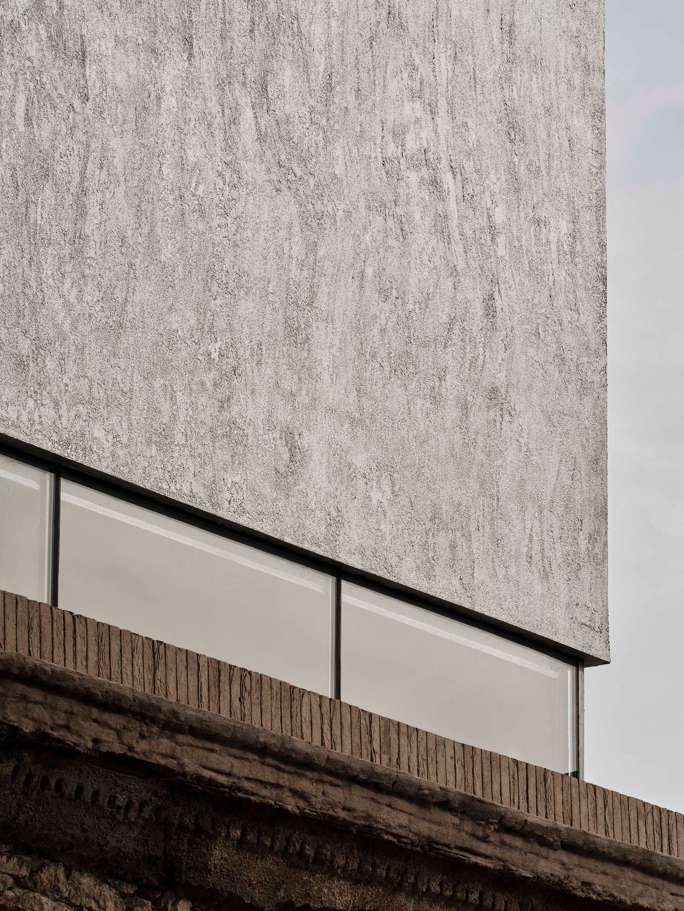 A close-up of the upper addition highlights its subtly mottled, rough-textured render, seemingly hovering above a ribbon of glazing. Below, vertically laid bricks and the eroded cornice of the original structure form a tactile transition between the contemporary volume and historic masonry. A close-up of the upper addition highlights its subtly mottled, rough-textured render, seemingly hovering above a ribbon of glazing. Below, vertically laid bricks and the eroded cornice of the original structure form a tactile transition between the contemporary volume and historic masonry.