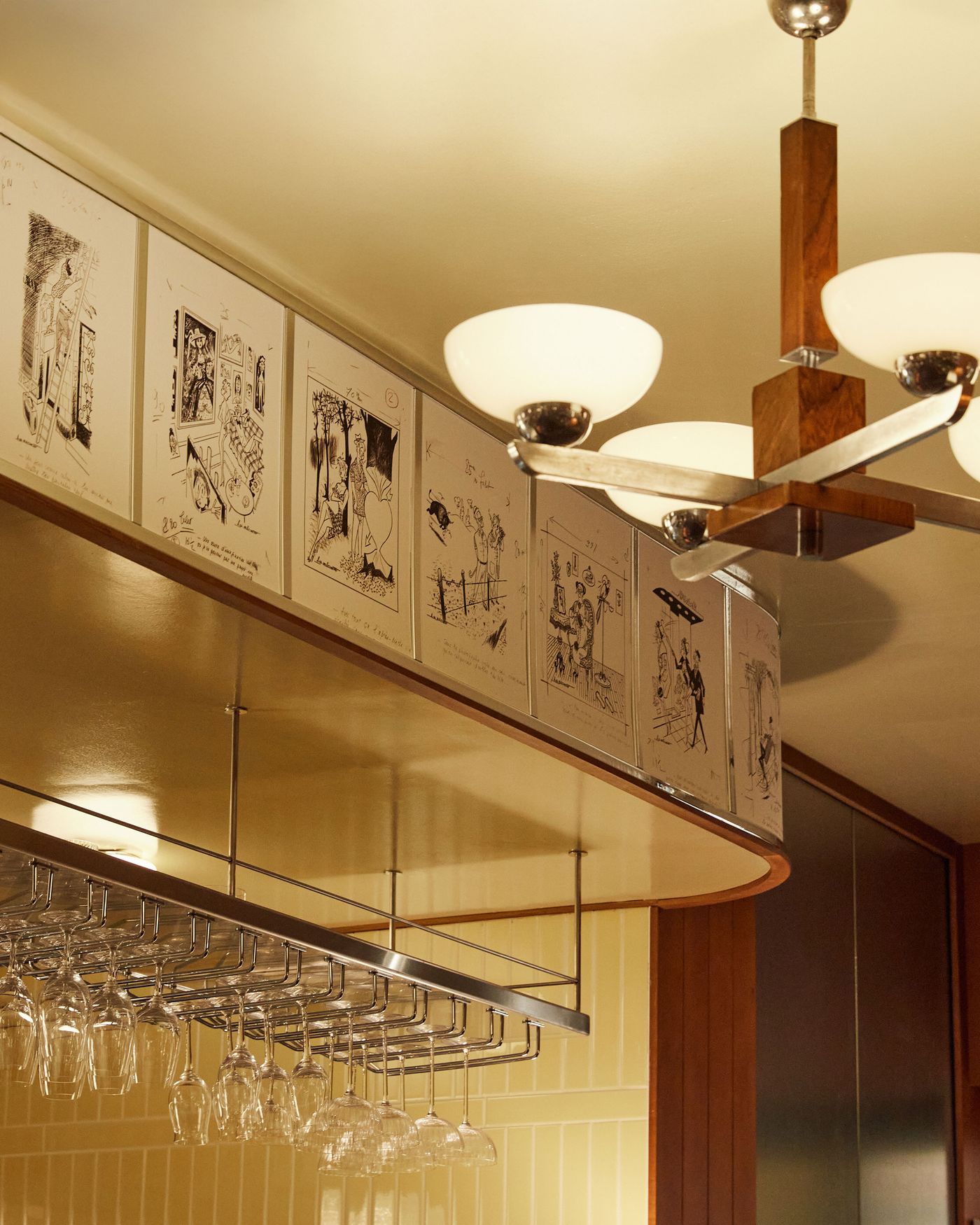 A close-up of a distinct mid-century modern light fixture, combining polished wood and metal with elegant, spherical glass shades. The fixture hangs in front of a curved frieze of framed black-and-white drawings. Below, a rack of hanging glassware and a wall of yellow tiles add to the space's rich and multi-layered aesthetic. A close-up of a distinct mid-century modern light fixture, combining polished wood and metal with elegant, spherical glass shades. The fixture hangs in front of a curved frieze of framed black-and-white drawings. Below, a rack of hanging glassware and a wall of yellow tiles add to the space's rich and multi-layered aesthetic.