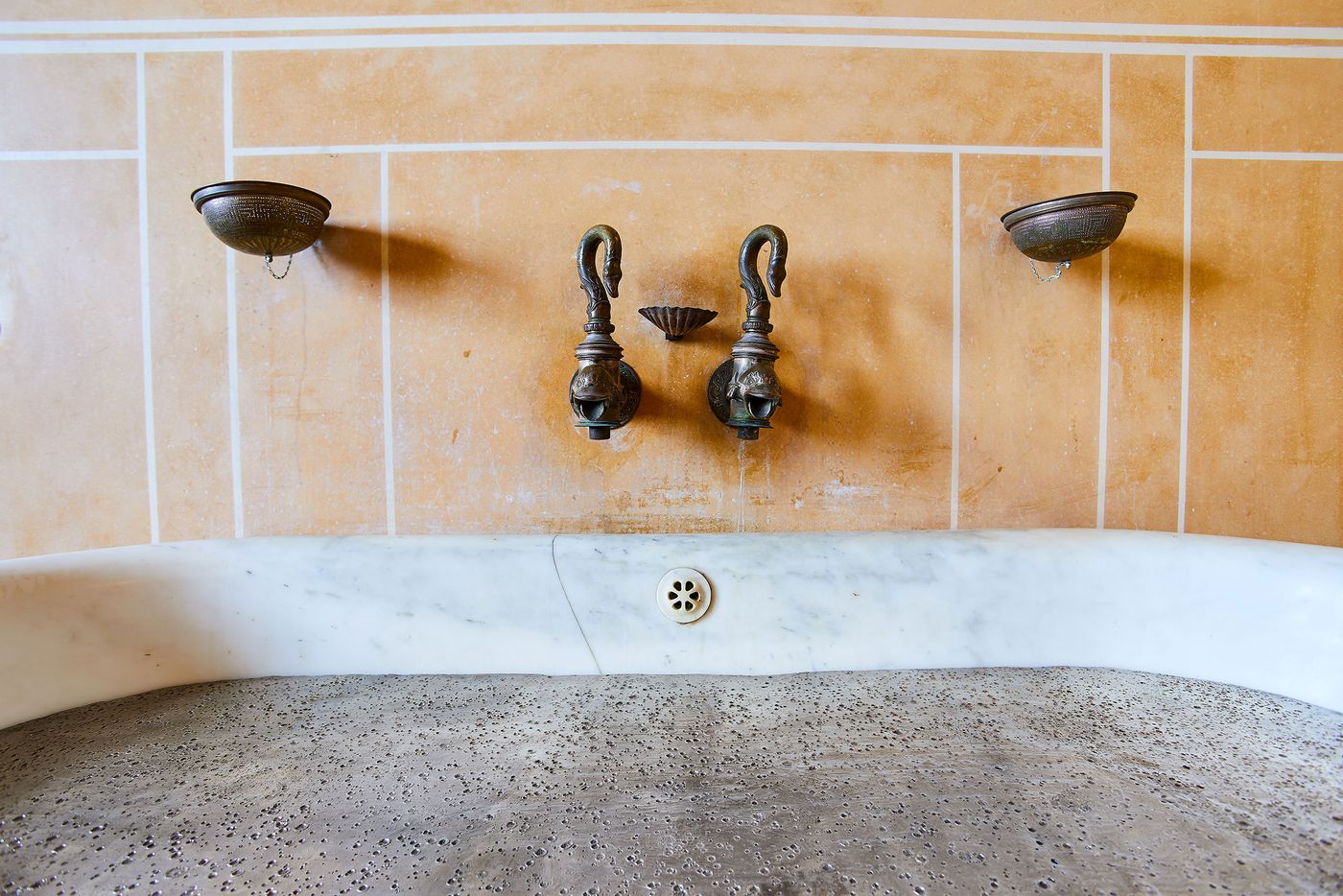 A close-up view of a classical bathing fixture. Two bronze faucets with swan heads sit on a pale orange wall, flanked by two small, bowl-shaped sconces. A thick, curved section of white marble forms the lip of the tub, which is filled with a dark, bronze surface, creating a stark visual and textural contrast. A close-up view of a classical bathing fixture. Two bronze faucets with swan heads sit on a pale orange wall, flanked by two small, bowl-shaped sconces. A thick, curved section of white marble forms the lip of the tub, which is filled with a dark, bronze surface, creating a stark visual and textural contrast.