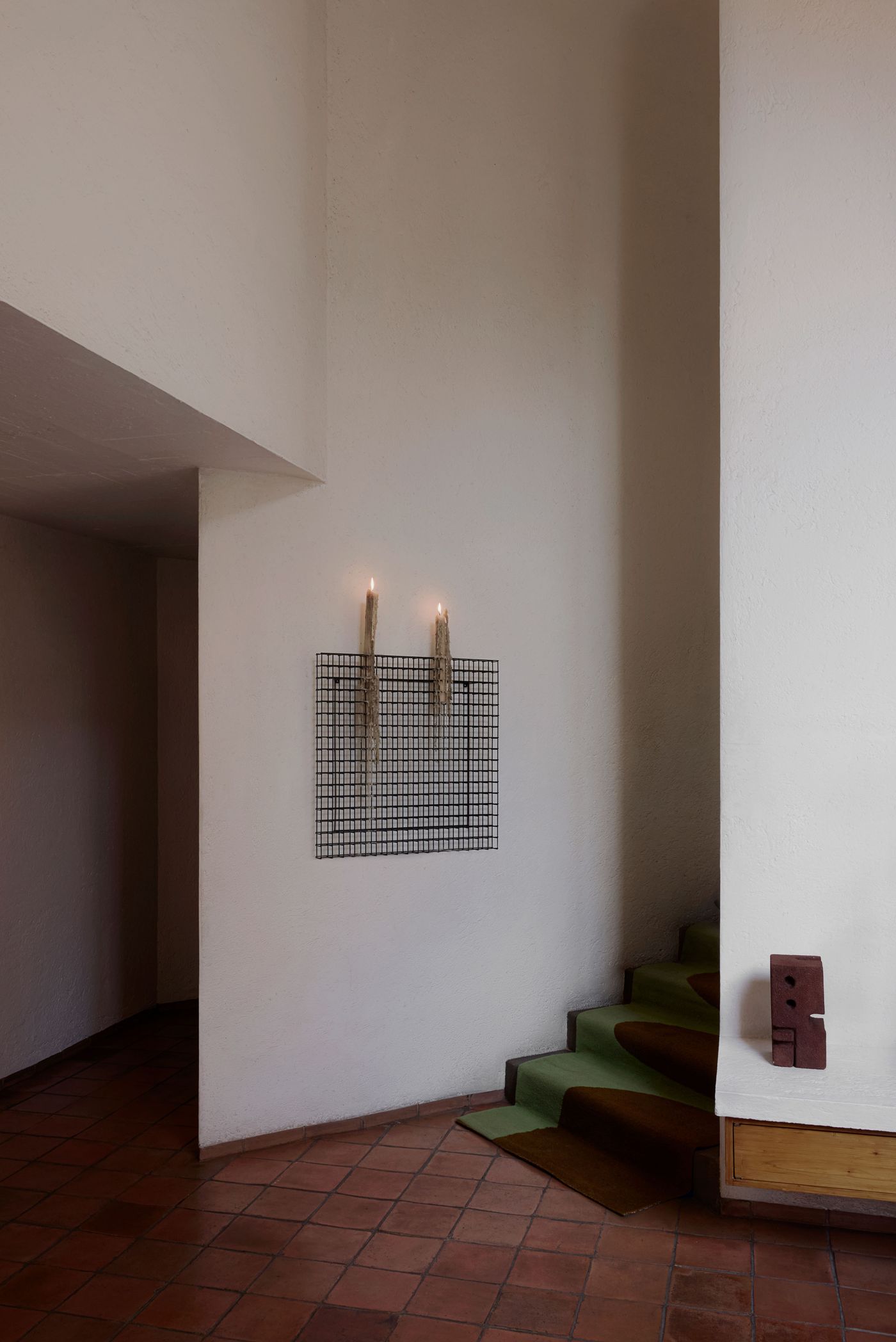 A minimalist interior corner with white walls, a grid candle holder, and a green and brown staircase on terracotta tiles.