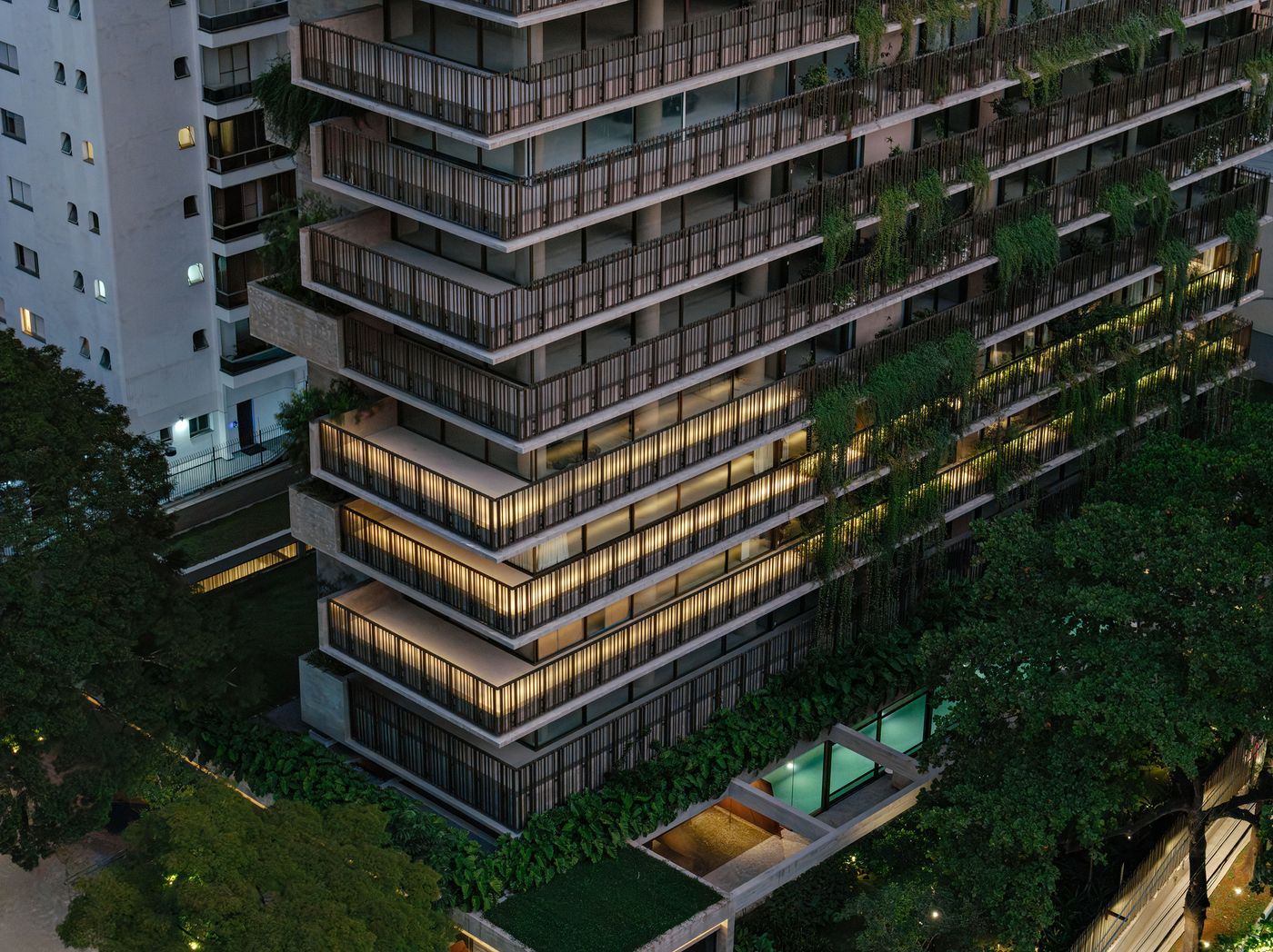 An evocative evening shot, showcasing its illuminated balconies and integrated vertical gardens that glow amidst the dimming light. The building's geometric concrete structure, softened by lush foliage, extends downwards to reveal a tranquil ground-level pool and landscaped areas, creating a harmonious urban oasis. An evocative evening shot, showcasing its illuminated balconies and integrated vertical gardens that glow amidst the dimming light. The building's geometric concrete structure, softened by lush foliage, extends downwards to reveal a tranquil ground-level pool and landscaped areas, creating a harmonious urban oasis.