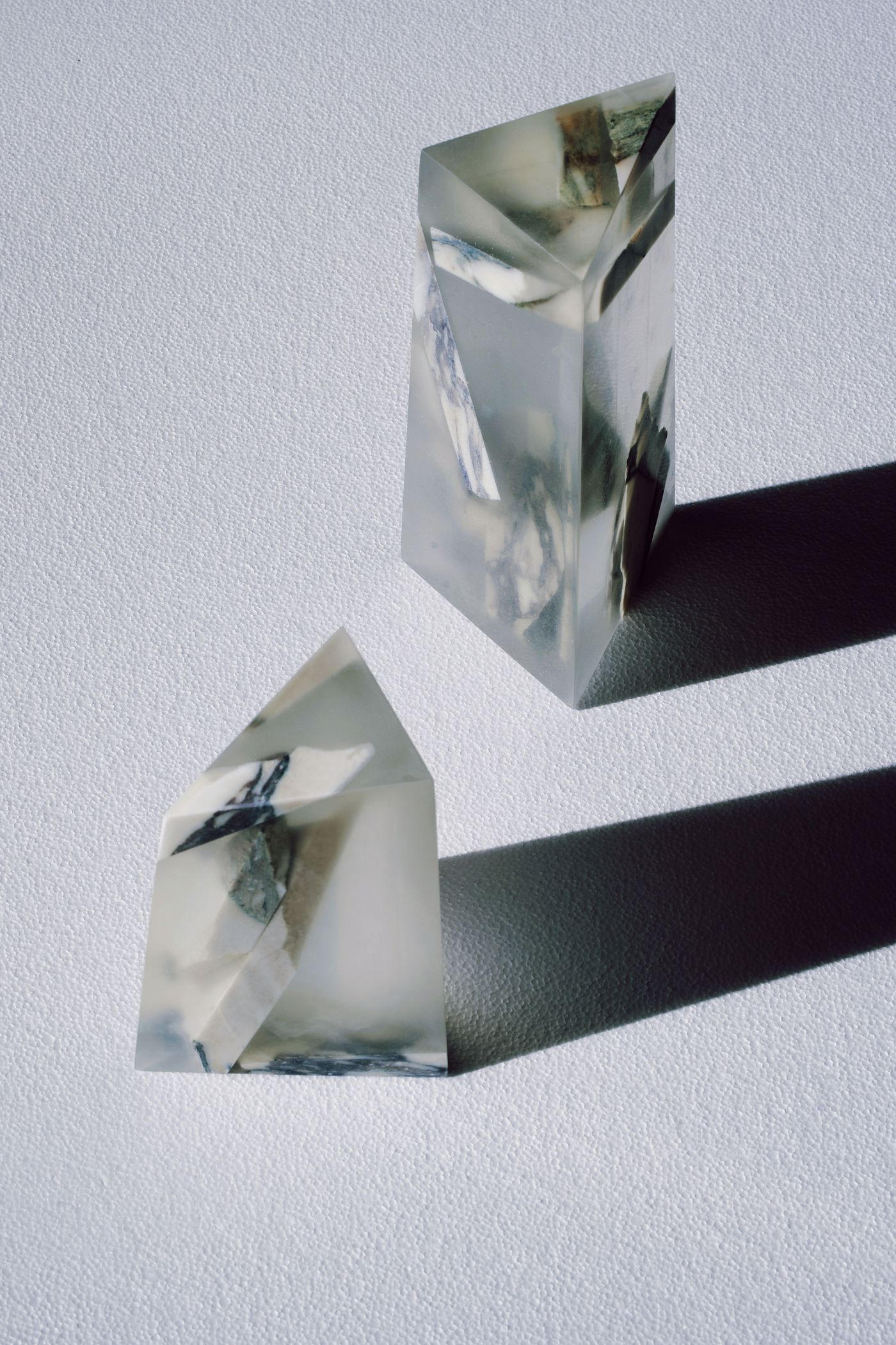 Two abstract geometric objects, resembling crystals or prisms, are presented against a neutral background. They are crafted from clear resin embedding fragments of marbled stone and dark blue or black material. The sharp angles and internal textures create intriguing patterns of light and shadow, suggesting a minimalist and sculptural approach to design, possibly as decorative objects or architectural elements.