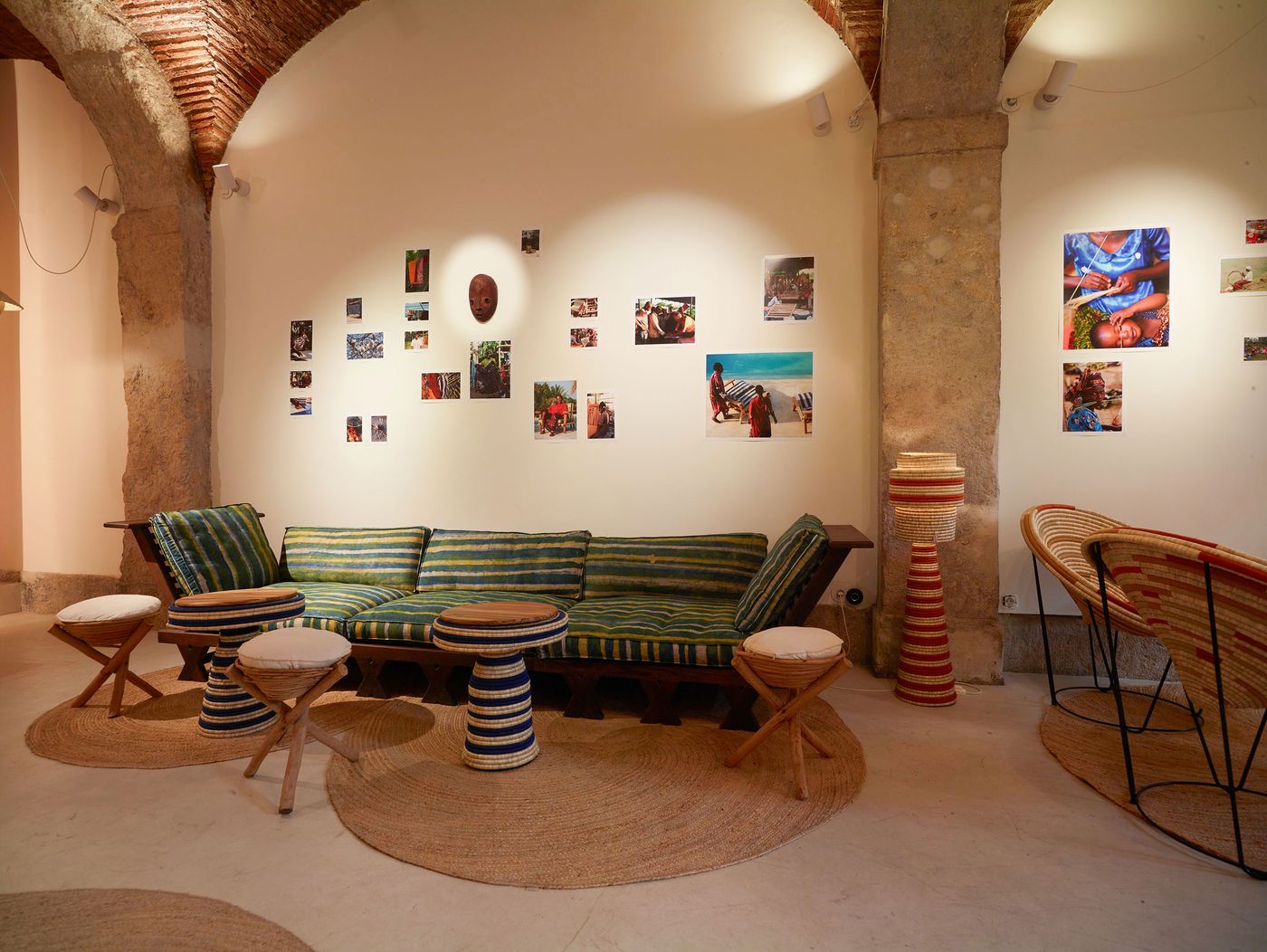 The "Kukua" exhibition at Prime Matter Gallery, presented during Lisbon Design Week, showcases a vibrant, bohemian-inspired seating area within an historic space. A low-slung wooden sofa with striped green and blue cushions is surrounded by natural fiber stools and round woven rugs. Exposed brick arches and stone columns frame walls adorned with diverse ethnographic photographs, creating an immersive, culturally rich atmosphere. The "Kukua" exhibition at Prime Matter Gallery, presented during Lisbon Design Week, showcases a vibrant, bohemian-inspired seating area within an historic space. A low-slung wooden sofa with striped green and blue cushions is surrounded by natural fiber stools and round woven rugs. Exposed brick arches and stone columns frame walls adorned with diverse ethnographic photographs, creating an immersive, culturally rich atmosphere.