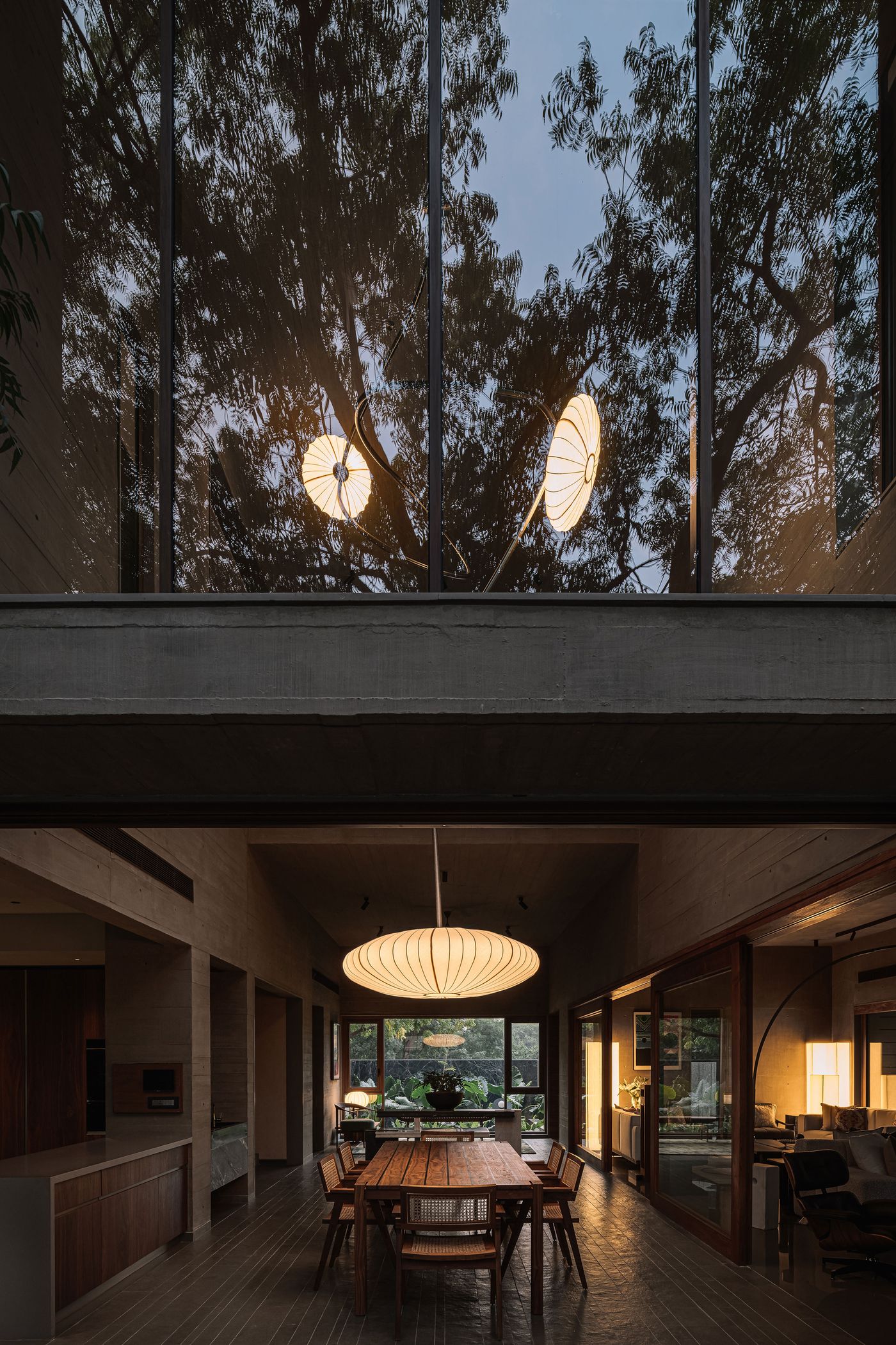 A captivating view into a sophisticated open-plan interior from a lower level, with a large, ribbed pendant light illuminating a long wooden dining table and rattan chairs. Above, expansive double-height windows reflect the silhouette of trees, interspersed with glowing abstract light fixtures, creating a dramatic interplay of light, architecture, and nature.