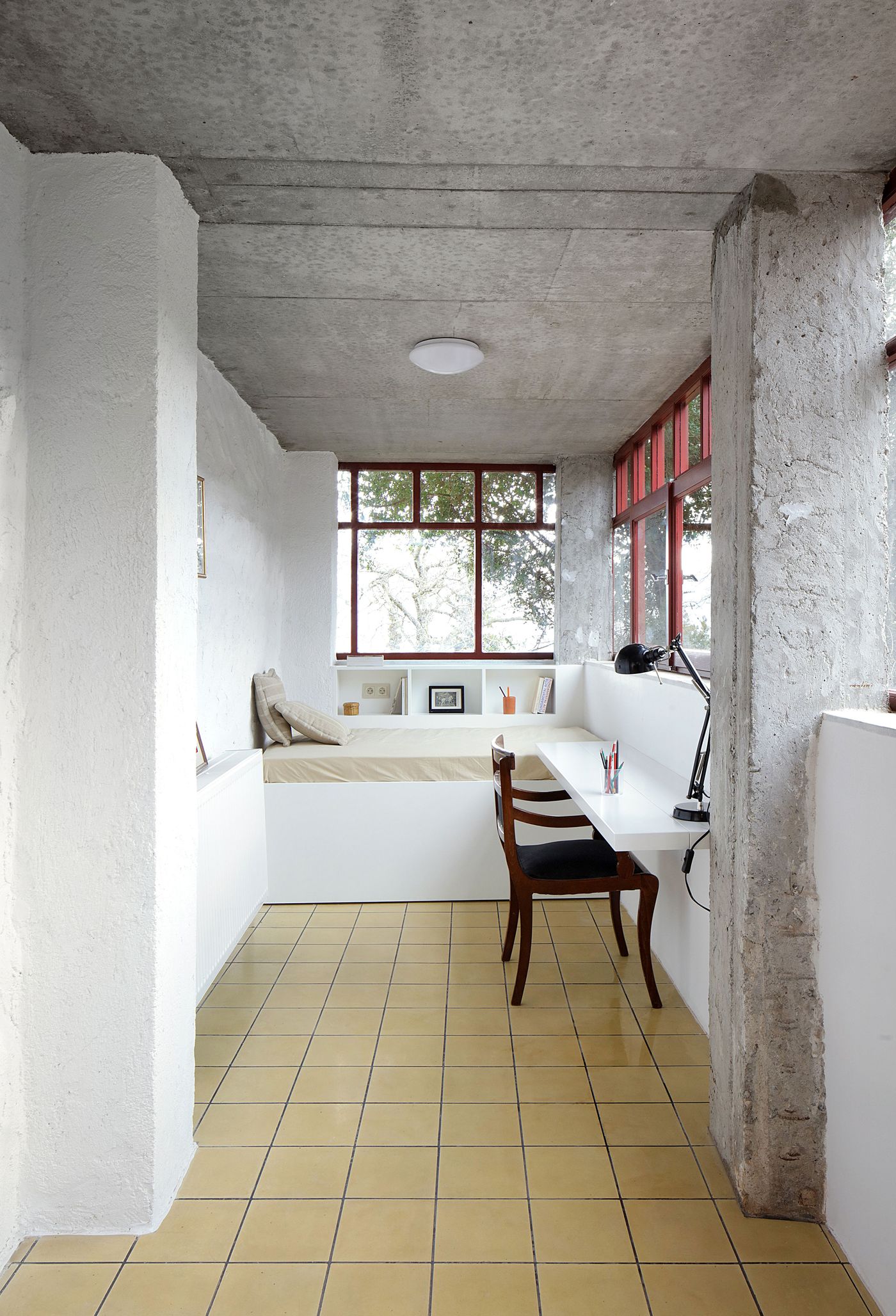 Yellow tile flooring extends throughout the compact study alcove, where exposed concrete structure creates honest material expression above the built-in seating platform. The minimalist intervention showcases how raw materials can be softened through careful color choices and thoughtful spatial proportions.