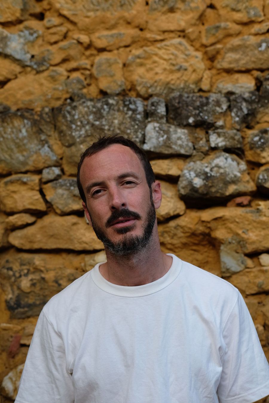 A man stands in front of a rustic stone wall, wearing a plain white t-shirt, exuding a calm and thoughtful presence. A man stands in front of a rustic stone wall, wearing a plain white t-shirt, exuding a calm and thoughtful presence.
