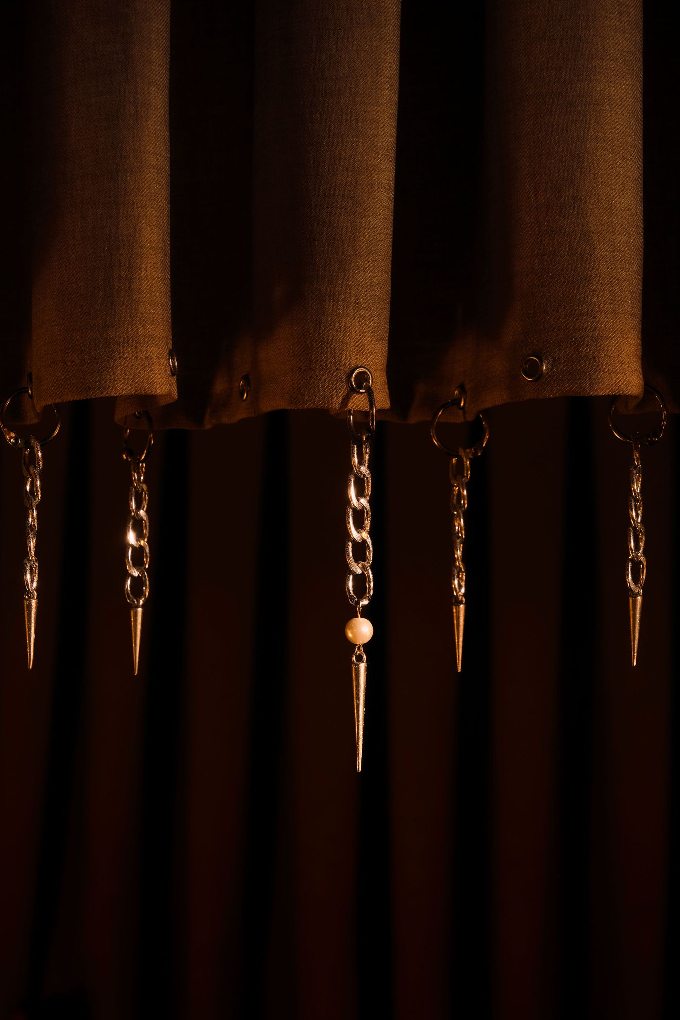 A close-up of the intriguing decorative elements at The Roses of Elagabalus: pointed, metallic "icicle" drops and small chains suspended from the bottom edge of a rich, dark fabric, likely a curtain or valance. The details are illuminated by a soft, warm light, emphasizing the luxurious and unique, slightly gothic yet opulent design philosophy.