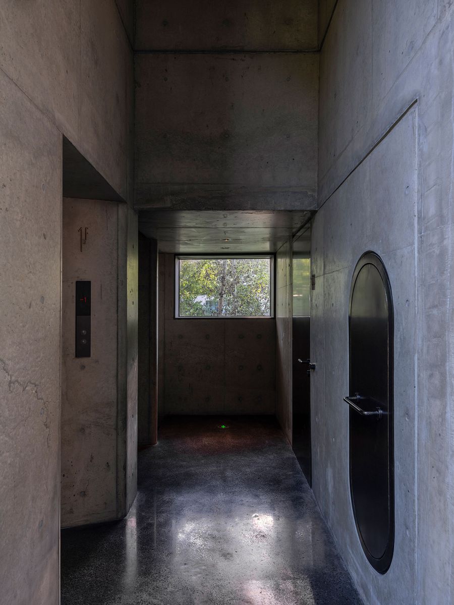 A minimalist corridor featuring concrete walls, a round black door, and an elevator, illuminated by natural light from a window. A minimalist corridor featuring concrete walls, a round black door, and an elevator, illuminated by natural light from a window.