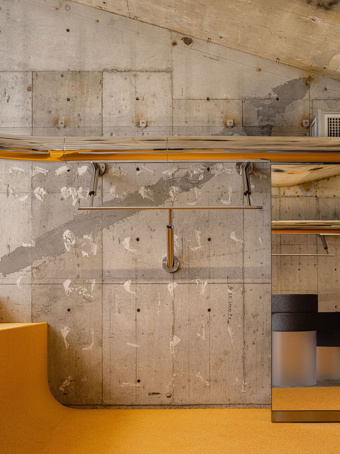 A close view of ANDERSSON BELL Tokyo’s upper floor pairs scarred Brutalist concrete with I IN’s polished chrome rail system and vivid yellow flooring. A mirrored panel extends the composition, while the curved junction between floor and display surface softens the otherwise hard-edged material palette.