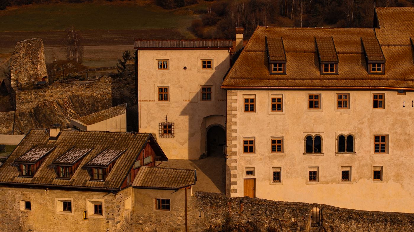Warm evening light falls on Castel Badia's layered façades: a gate tower with a vaulted passage, a main wing with painted quoins, twin-arched Romanesque windows and an ornate wrought-iron window grille. Shingled roofs with timber dormers, a rubble stone wall and a crisp contemporary addition complete the composition. Warm evening light falls on Castel Badia's layered façades: a gate tower with a vaulted passage, a main wing with painted quoins, twin-arched Romanesque windows and an ornate wrought-iron window grille. Shingled roofs with timber dormers, a rubble stone wall and a crisp contemporary addition complete the composition.