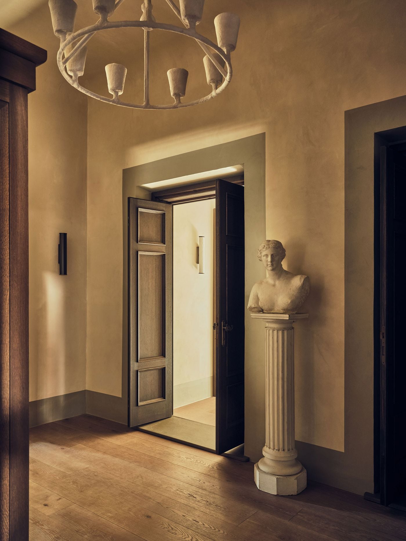 A classical female bust on a fluted column stands sentinel beside panelled oak doors left ajar on a landing at La Réserve Firenze. A ring chandelier of plaster cups, warm plastered walls, painted door surrounds and wide oak boards bathed in low light create a scene of theatrical stillness. A classical female bust on a fluted column stands sentinel beside panelled oak doors left ajar on a landing at La Réserve Firenze. A ring chandelier of plaster cups, warm plastered walls, painted door surrounds and wide oak boards bathed in low light create a scene of theatrical stillness.