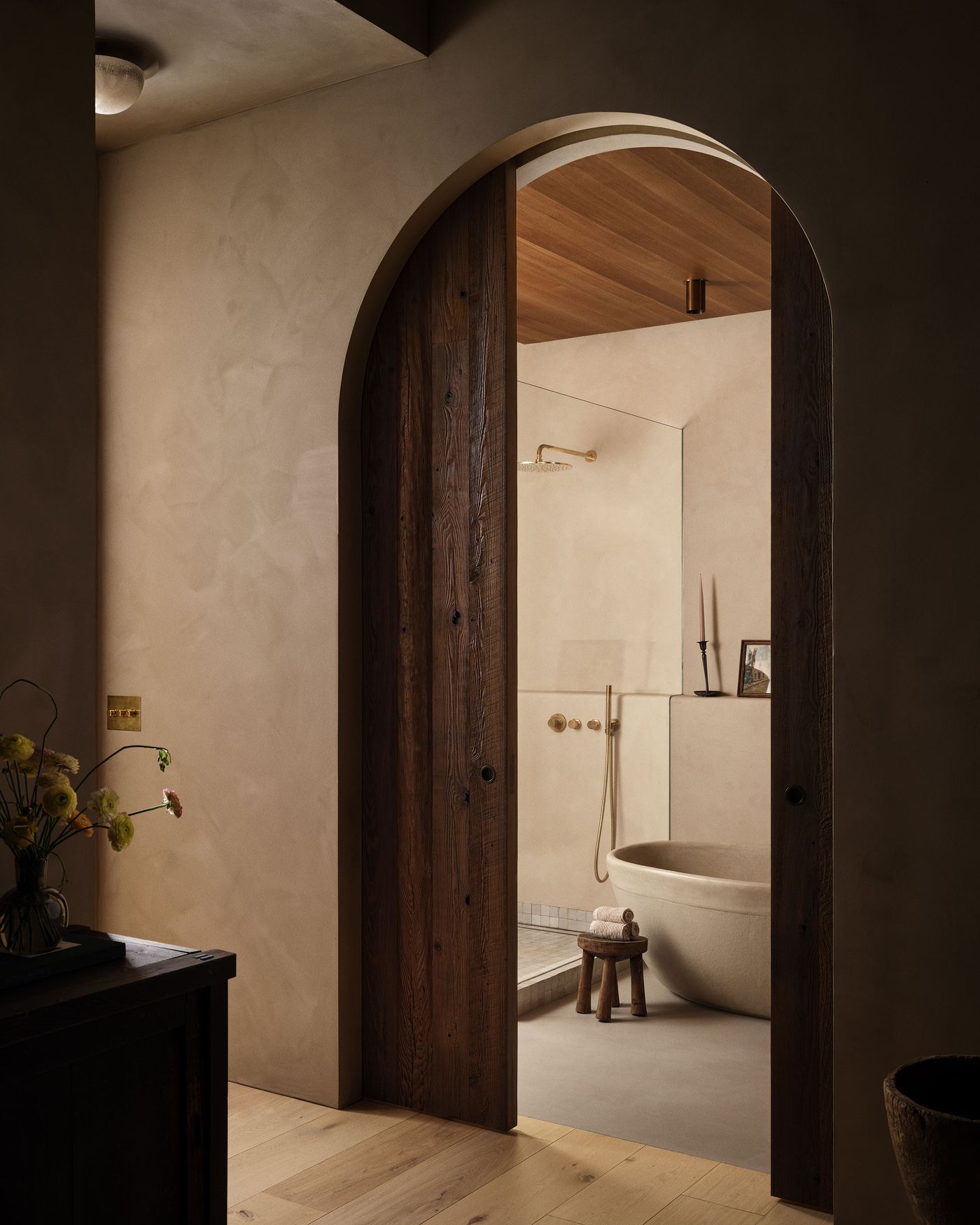 A serene bathroom view reveals a sleek tub, gold fixtures, and natural details, framed by an arched wooden doorway. Soft, warm lighting enhances the ambiance.