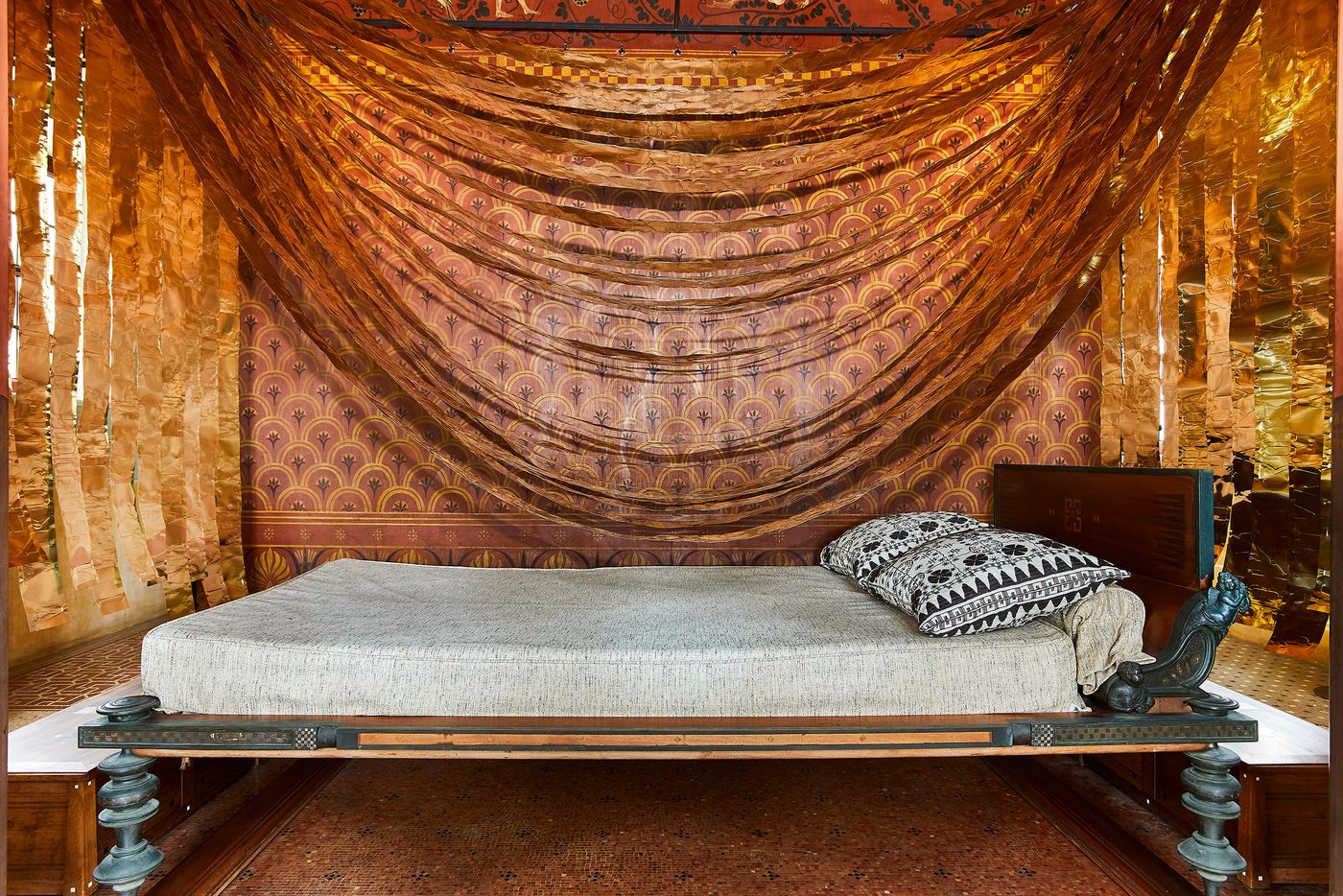 An opulent daybed, featuring carved wooden legs and a linen-colored mattress with throw pillows. The space is a striking blend of ancient and modern design, with a wall mural in red and gold patterns. A dramatic canopy of billowing, sheer fabric in a vibrant orange hangs from above, flanked by panels of a crinkled golden foil installation by Gabriel Leger. An opulent daybed, featuring carved wooden legs and a linen-colored mattress with throw pillows. The space is a striking blend of ancient and modern design, with a wall mural in red and gold patterns. A dramatic canopy of billowing, sheer fabric in a vibrant orange hangs from above, flanked by panels of a crinkled golden foil installation by Gabriel Leger.