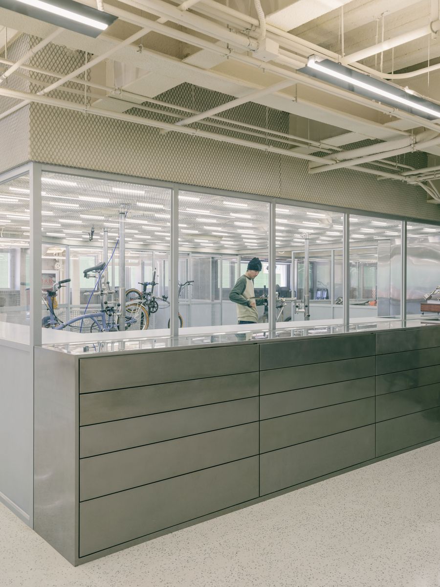 A meticulously designed bicycle workshop within AITASHOP is enclosed by transparent glass and perforated metal panels, allowing visitors to observe the craft. A long, dark grey storage unit acts as a counter in the foreground, while a technician works on a bike, highlighting the shop's commitment to both display and service in a clean, industrial-minimalist setting. A meticulously designed bicycle workshop within AITASHOP is enclosed by transparent glass and perforated metal panels, allowing visitors to observe the craft. A long, dark grey storage unit acts as a counter in the foreground, while a technician works on a bike, highlighting the shop's commitment to both display and service in a clean, industrial-minimalist setting.