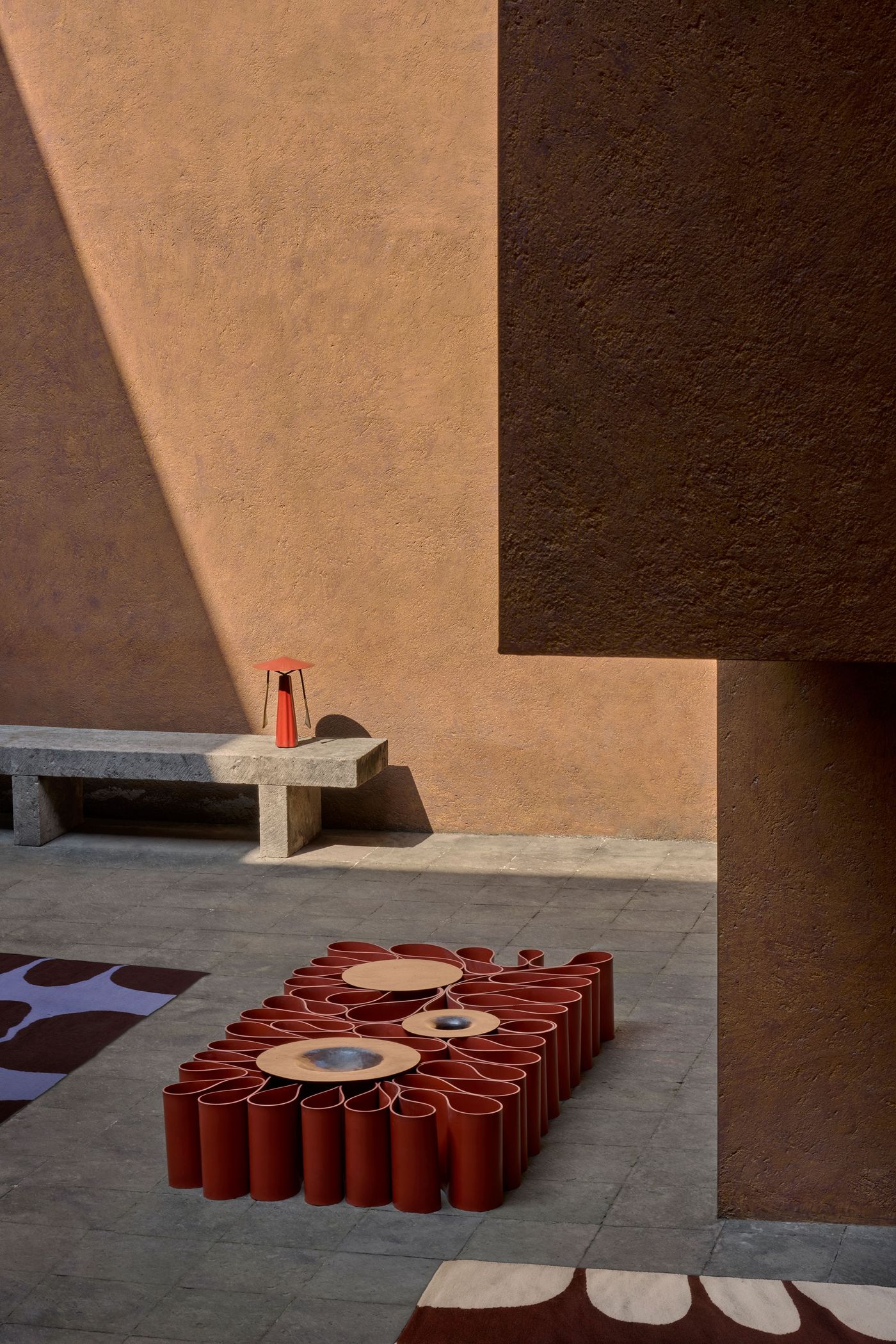 A modern outdoor space featuring a unique red and beige sculptural table, grey stone bench, and textured brown walls. Natural light creates shadows.