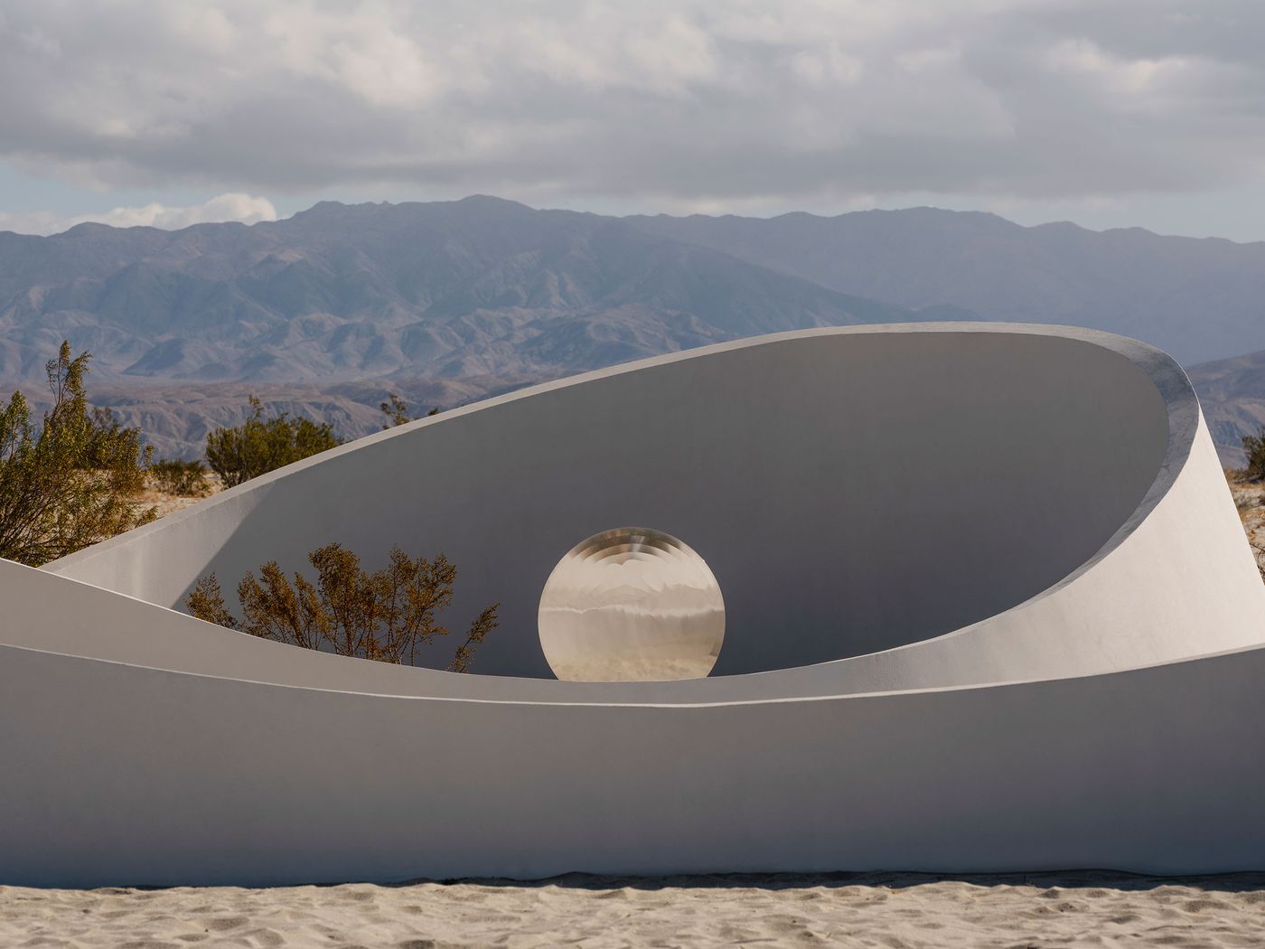 A modern, curved white sculpture encloses a shiny sphere, set against a backdrop of distant mountains and cloudy sky.