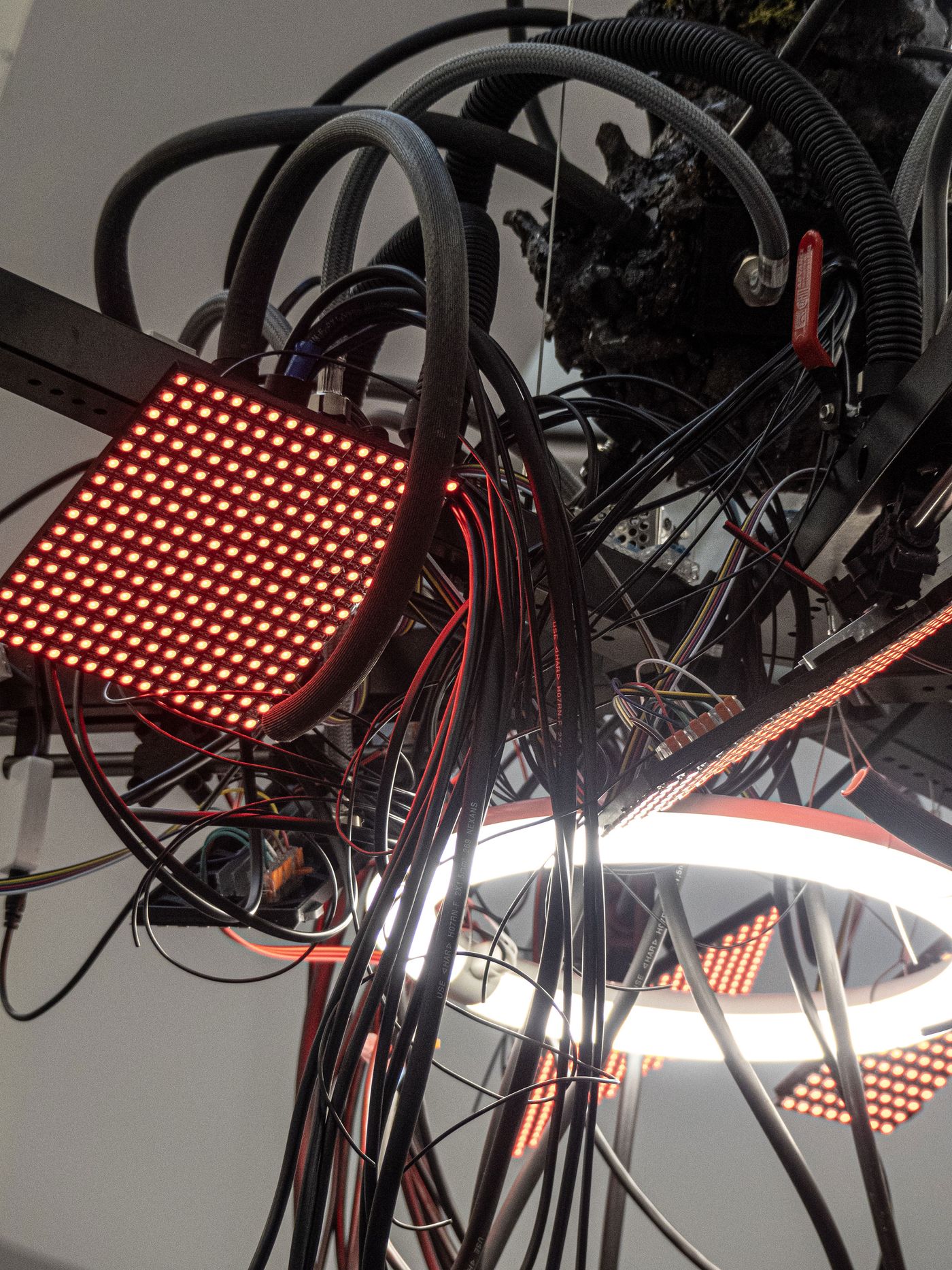 A tangled arrangement of black and colored wires with red LED panels and a circular white light, suspended from a structure.