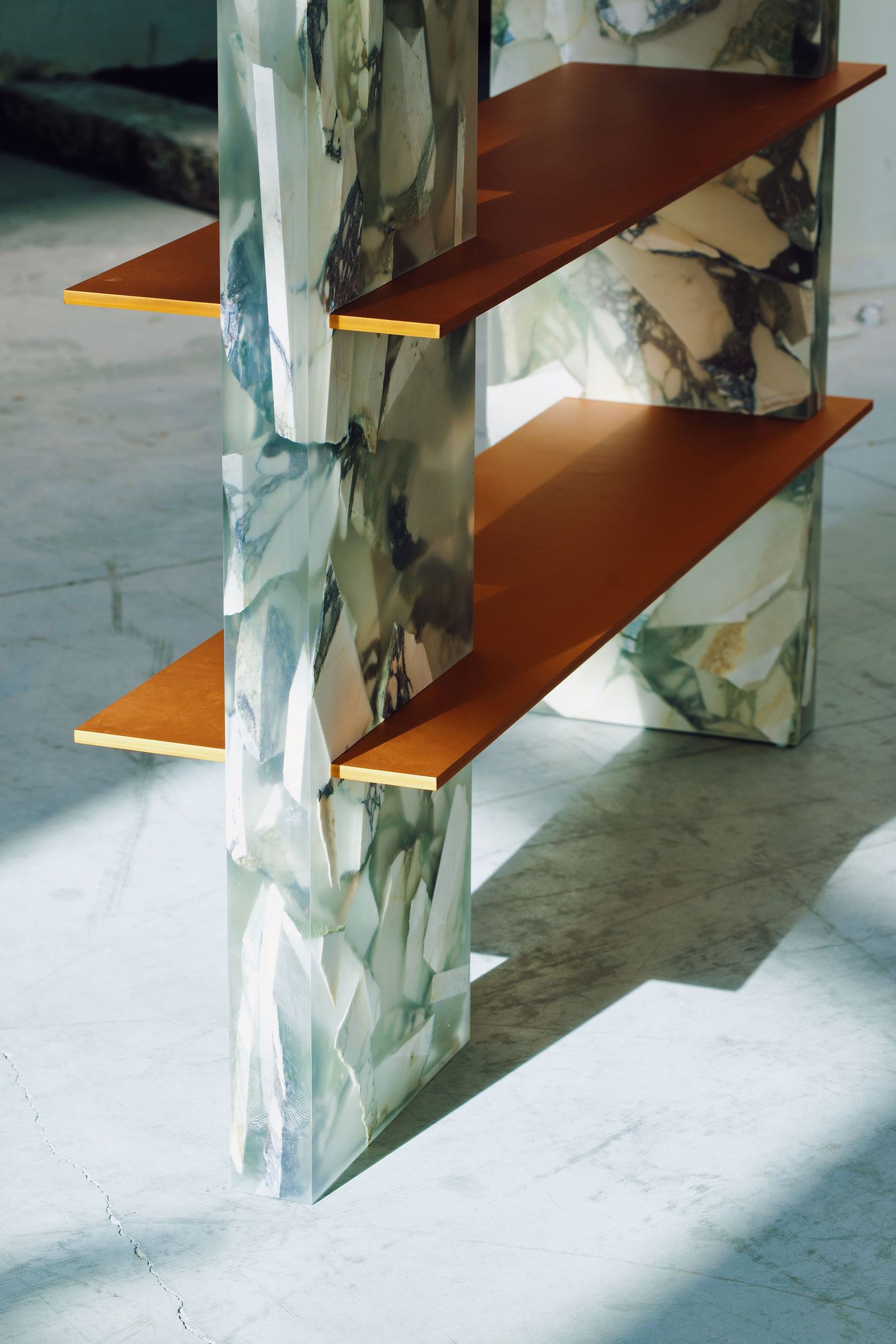 A close-up view of a contemporary shelving unit reveals its unique construction. The vertical supports are made of stacked, clear resin blocks containing fragments of marbled stone, creating a visually complex and translucent effect. Thin, warm-toned shelves slot into these supports, contrasting with the intricate patterns of the resin. The play of light and shadow on the polished floor adds depth to the composition.