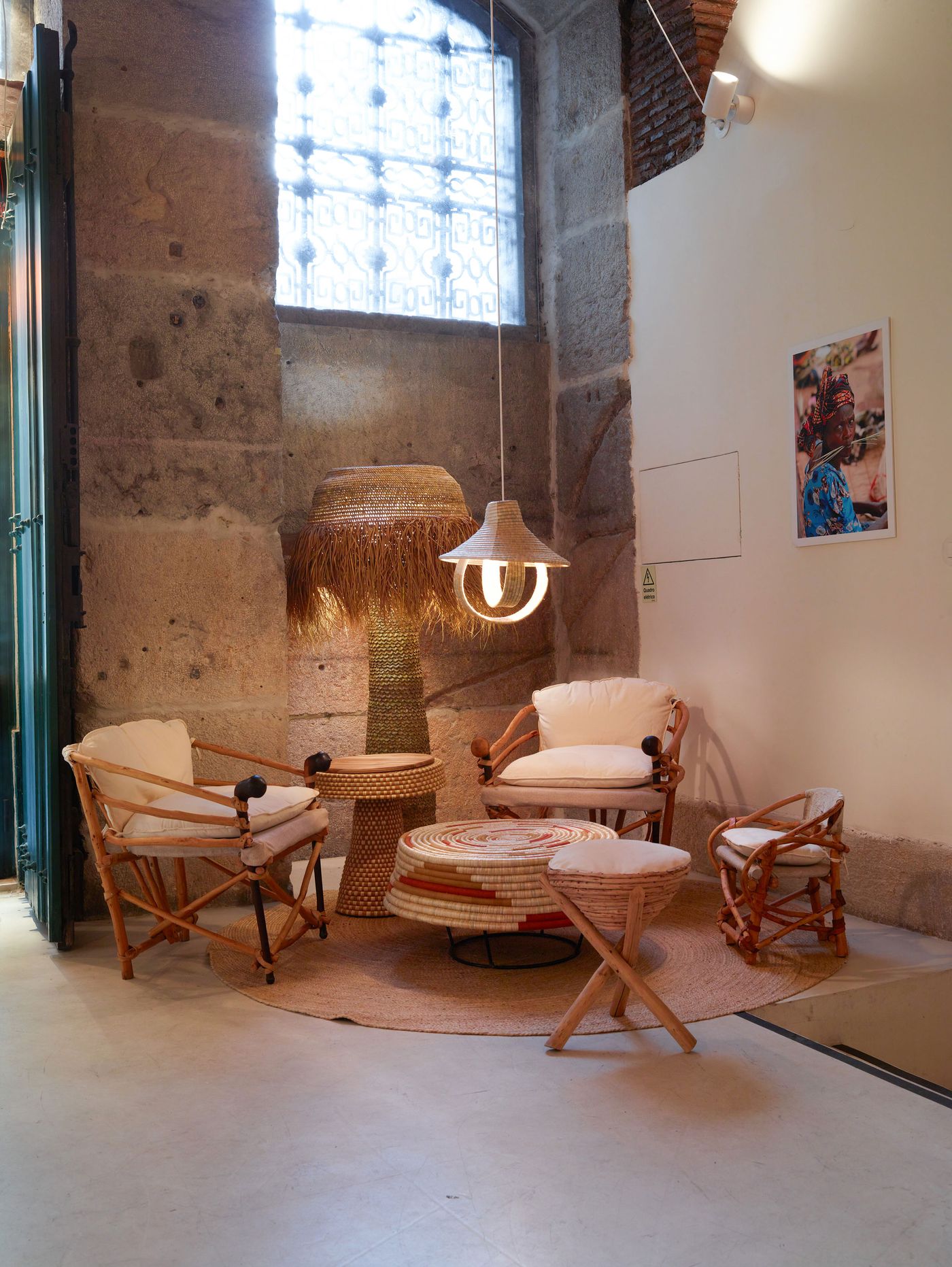 A seating vignette within the "Kukua" exhibition at Prime Matter Gallery, featuring an eclectic mix of natural materials. Two light-toned rattan chairs with cream cushions frame a circular woven coffee table. Overhead, two unique pendant lights, one with a fringed straw shade and another with a woven, sculptural form, cast warm light on the stone walls and concrete floor, creating an inviting, artisanal ambiance.