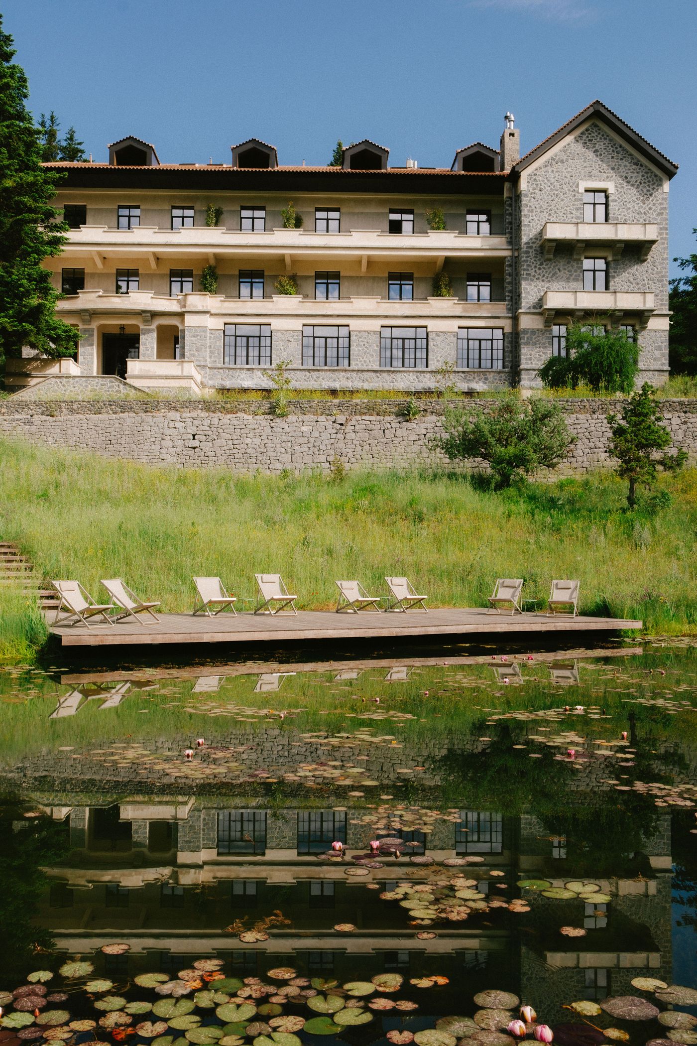 A grand neoclassical hotel, with its facade reflected in a calm pond dotted with water lilies, stands above a tiered stone wall and lush green lawn. A wooden deck on the pond's edge holds several light-colored lounge chairs, offering a serene outdoor seating area that blends historical grandeur with natural tranquility.