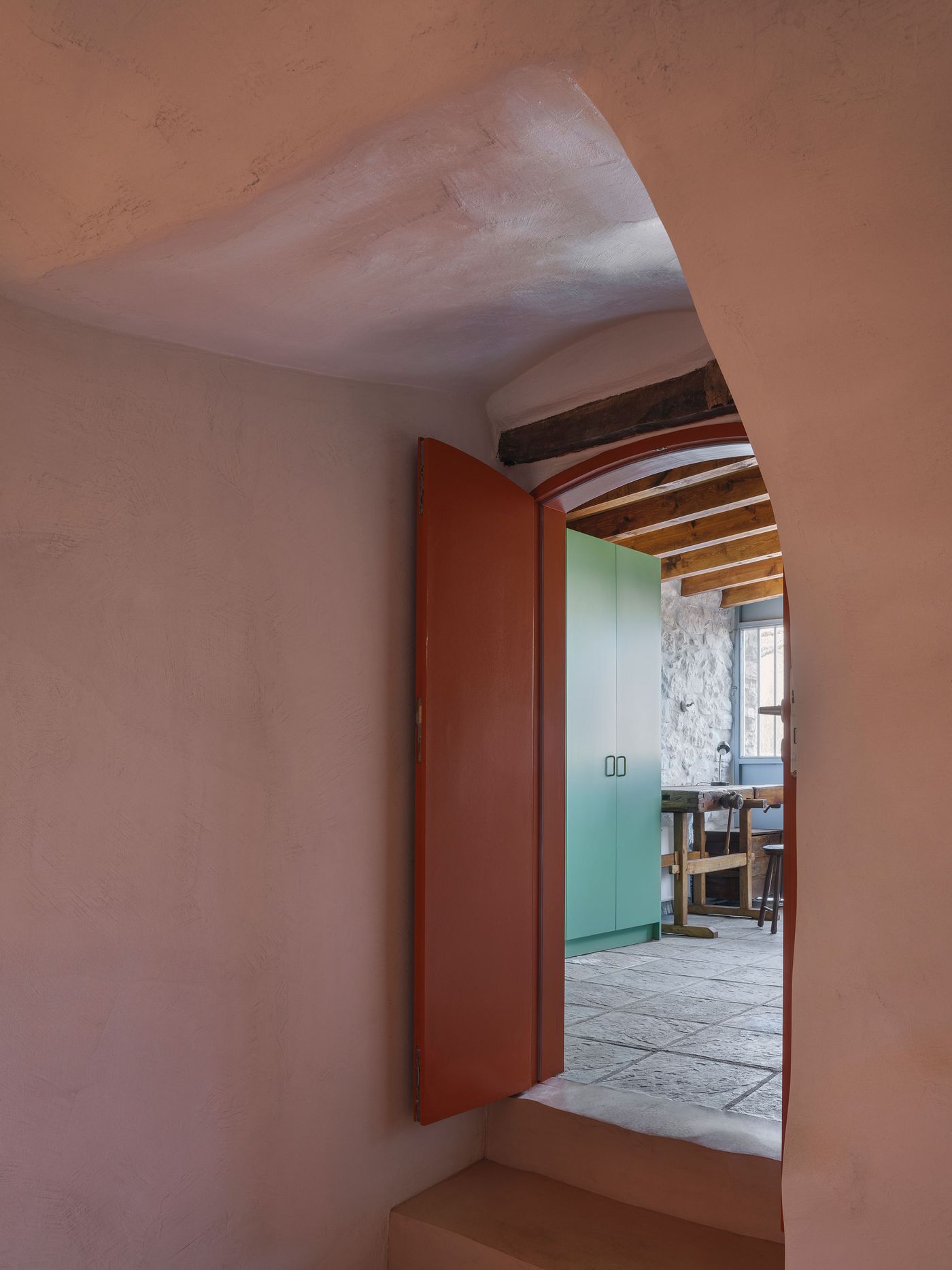 Warm coral-toned walls frame an arched passage leading to the communal areas, where exposed timber roof structure creates dramatic geometric patterns. The soft color palette provides intimate contrast to the robust stone construction, demonstrating how contemporary restoration can enhance rather than compete with historical architecture.
