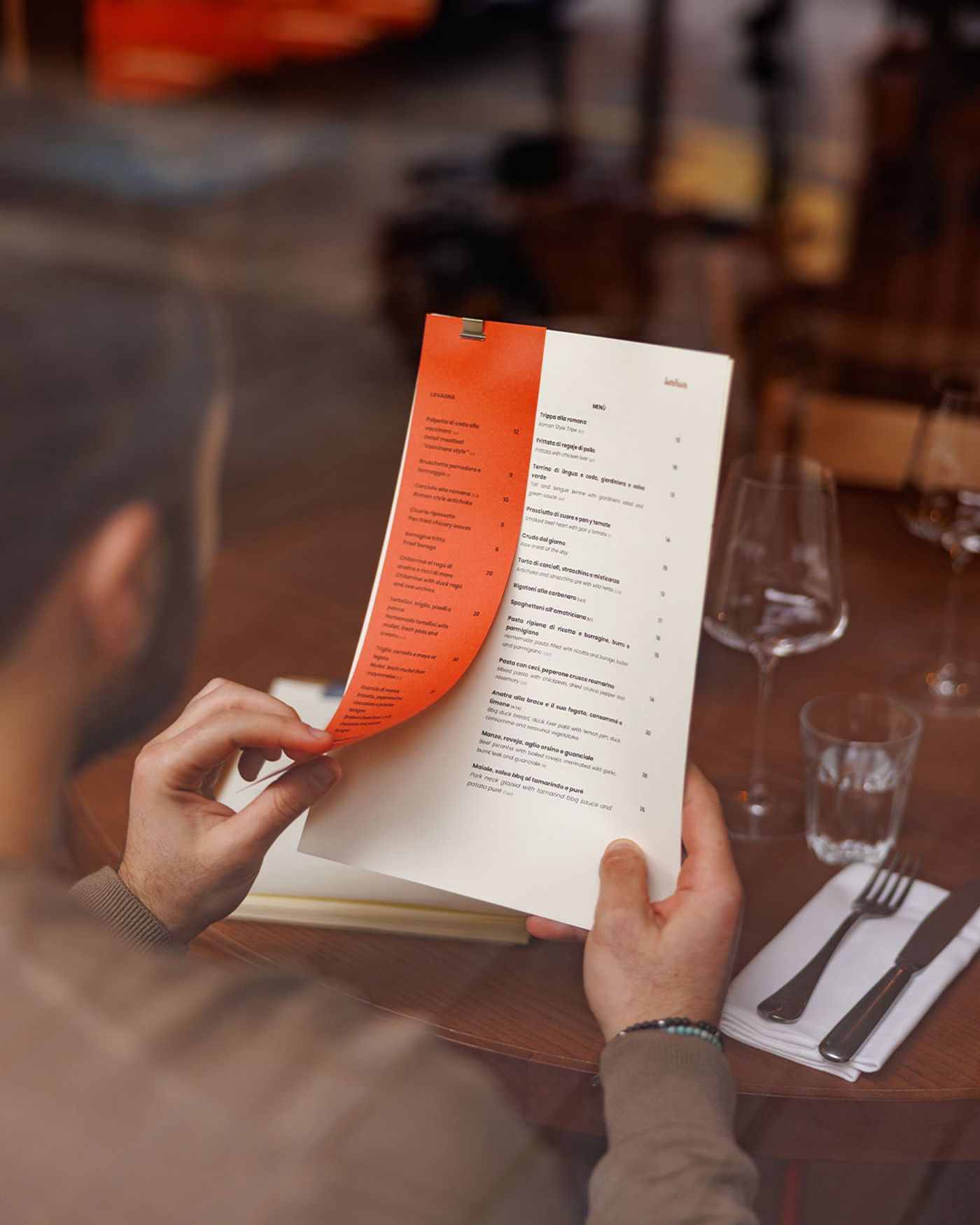 A person's hands delicately hold an open menu at Santo Palato, revealing a list of Italian dishes. The warm lighting highlights the menu's orange spine, with a glass of water and cutlery on a white napkin positioned to the right, suggesting an immersive and focused dining experience.