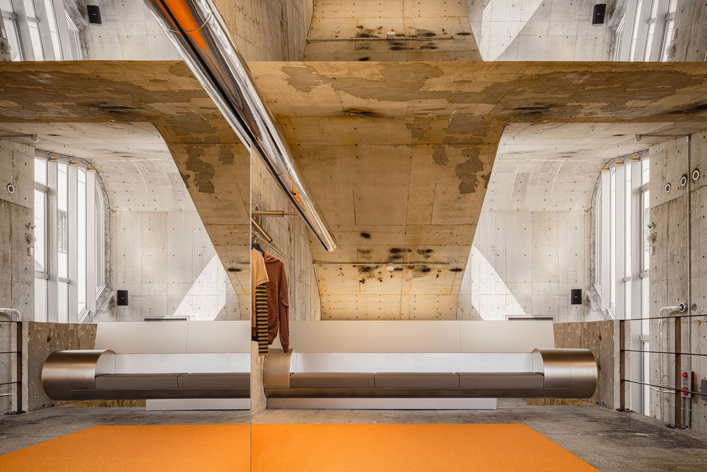 Andersson Bell Tokyo flagship by I IN: a full-height mirror doubles the upper floor into a near-symmetrical composition, an elongated bench capped with rounded brushed-steel ends floating above orange microcement, set beneath a soaring board-marked concrete shell scored by sloping beams, tall windows and a mirror-polished steel rail.