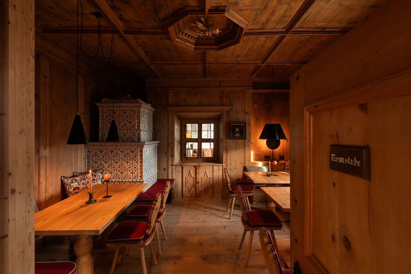 A panelled dining parlour in knotty pine, is crowned by a coffered ceiling with an octagonal rosette and gilded carving. A blue-patterned tiled stove, black conical pendants, solid tables, carved chairs with red cushions and a small interior window into the adjoining lounge complete the room. A panelled dining parlour in knotty pine, is crowned by a coffered ceiling with an octagonal rosette and gilded carving. A blue-patterned tiled stove, black conical pendants, solid tables, carved chairs with red cushions and a small interior window into the adjoining lounge complete the room.
