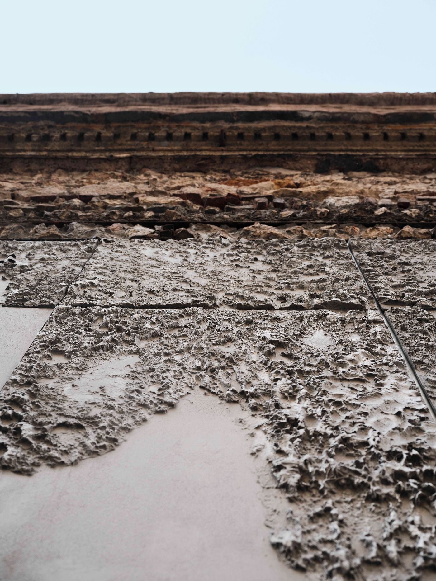 Seen from below, the entrance door’s textured metal panels echo the rugged stonework and eroded brick detailing of the historic façade above. Smooth sections interrupt the heavily reliefed surfaces, creating an abstract patchwork that reinterprets the irregular materiality of the original building. Seen from below, the entrance door’s textured metal panels echo the rugged stonework and eroded brick detailing of the historic façade above. Smooth sections interrupt the heavily reliefed surfaces, creating an abstract patchwork that reinterprets the irregular materiality of the original building.