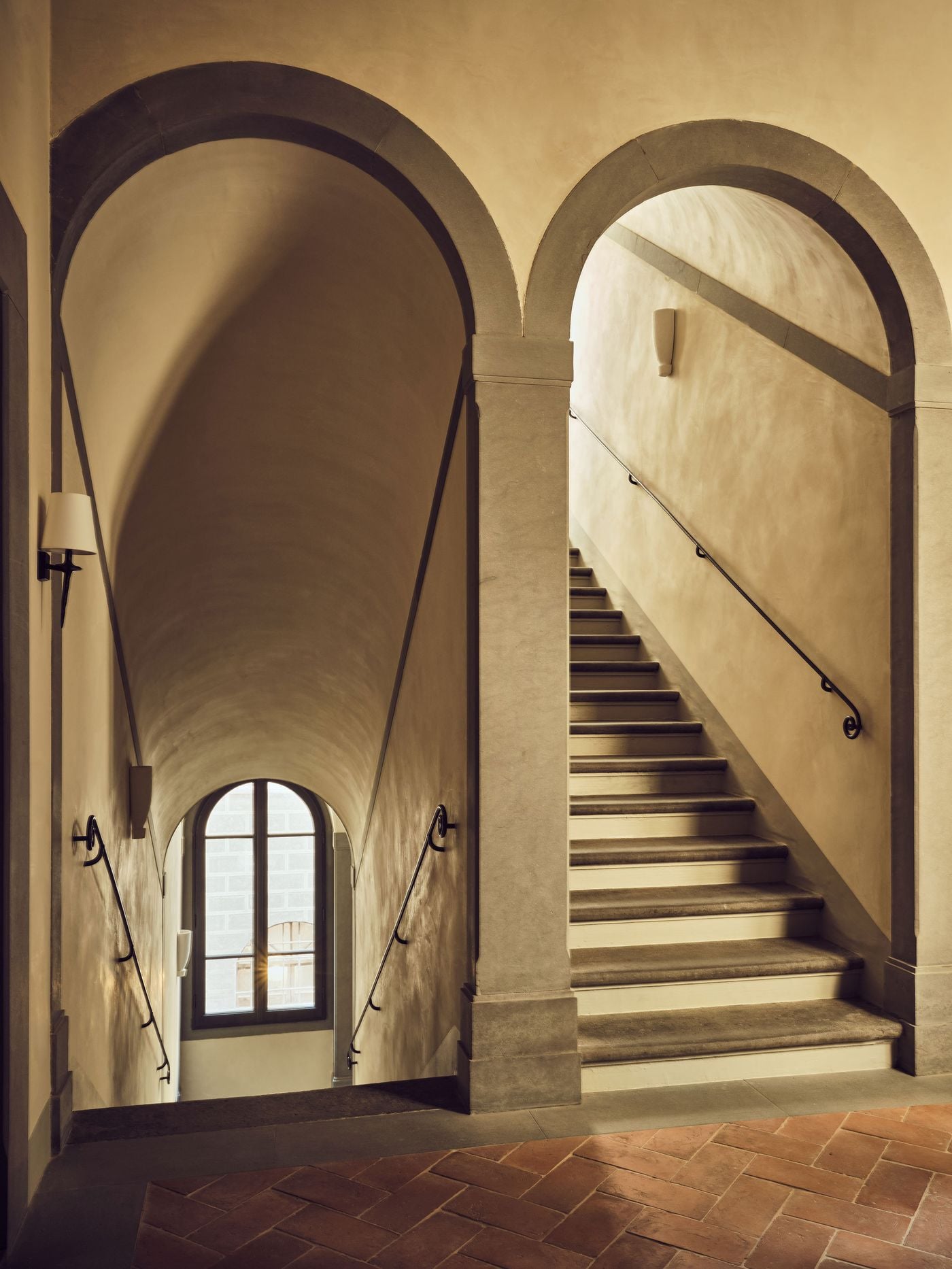 Twin arches framed in grey stone divide the stairwell at La Réserve Firenze: one descends beneath a barrel vault towards a light-filled arched window, the other climbs a flight of worn stone steps. Scrolled iron handrails, plaster sconces and a herringbone terracotta floor articulate the palazzo's original Renaissance architecture. Twin arches framed in grey stone divide the stairwell at La Réserve Firenze: one descends beneath a barrel vault towards a light-filled arched window, the other climbs a flight of worn stone steps. Scrolled iron handrails, plaster sconces and a herringbone terracotta floor articulate the palazzo's original Renaissance architecture.