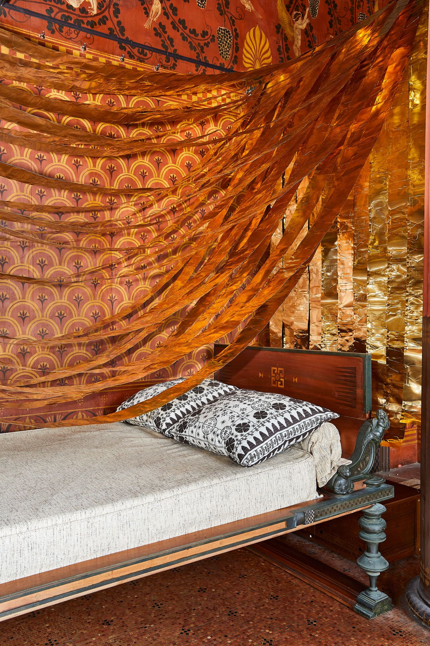 A detail shot of the daybed area. A sheer, rust-colored fabric billows elegantly above the bed, contrasting with the detailed, patterned wall mural. The head of the bed is a dark wood panel with an engraved pattern, and two pillows in a contrasting pattern rest on the bed, adding to the eclectic and richly layered aesthetic.