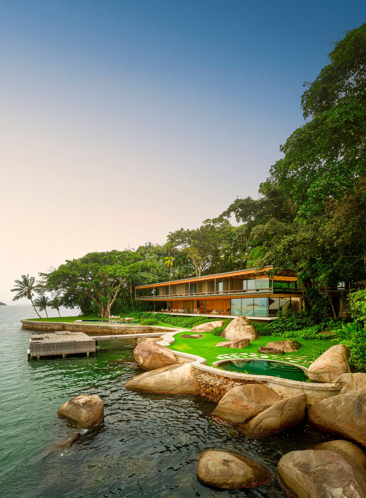 A wide shot captures the modernist dwelling nestled into the lush Brazilian coastline, its horizontal form and warm timber accents mirroring the natural contours. Terraced lawns, natural rock formations, and organic pools lead down to a private dock, creating a sophisticated yet utterly connected biophilic retreat on the tranquil emerald sea.
