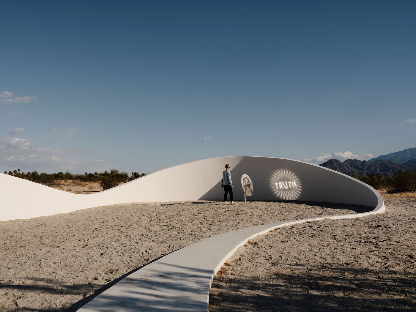 A person stands on sandy terrain beside a curved white wall, illuminated with the word "TRUTH" against a clear blue sky.