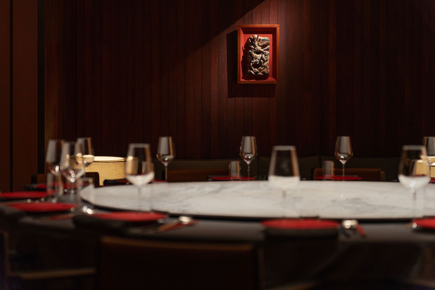 Elegant restaurant setting with a round marble table, wine glasses, red-topped napkins, and a decorative wooden artwork on the wall. Elegant restaurant setting with a round marble table, wine glasses, red-topped napkins, and a decorative wooden artwork on the wall.