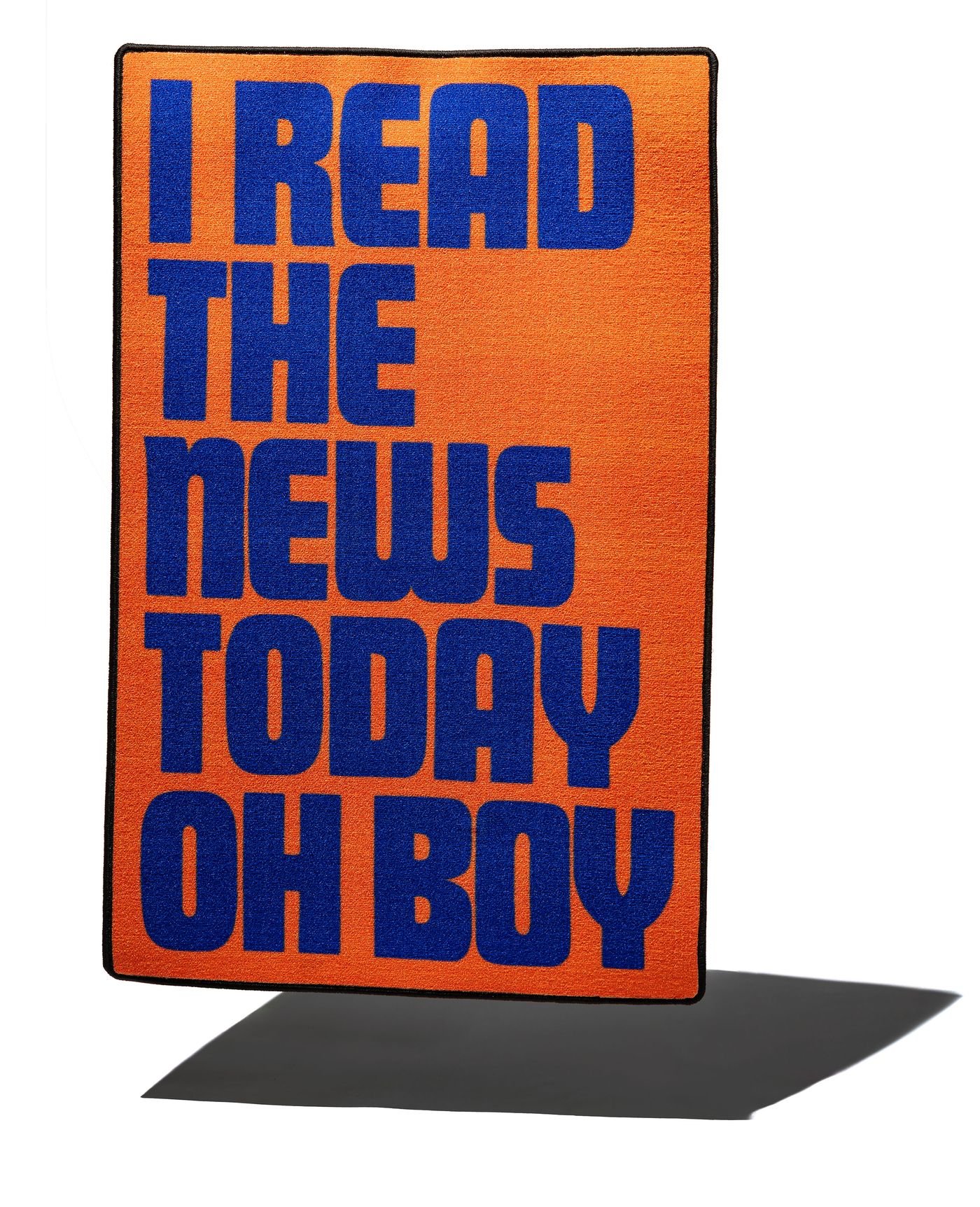 Bright orange doormat with bold blue text reading "I READ THE NEWS TODAY OH BOY." The mat has a black border and a shadow beneath.