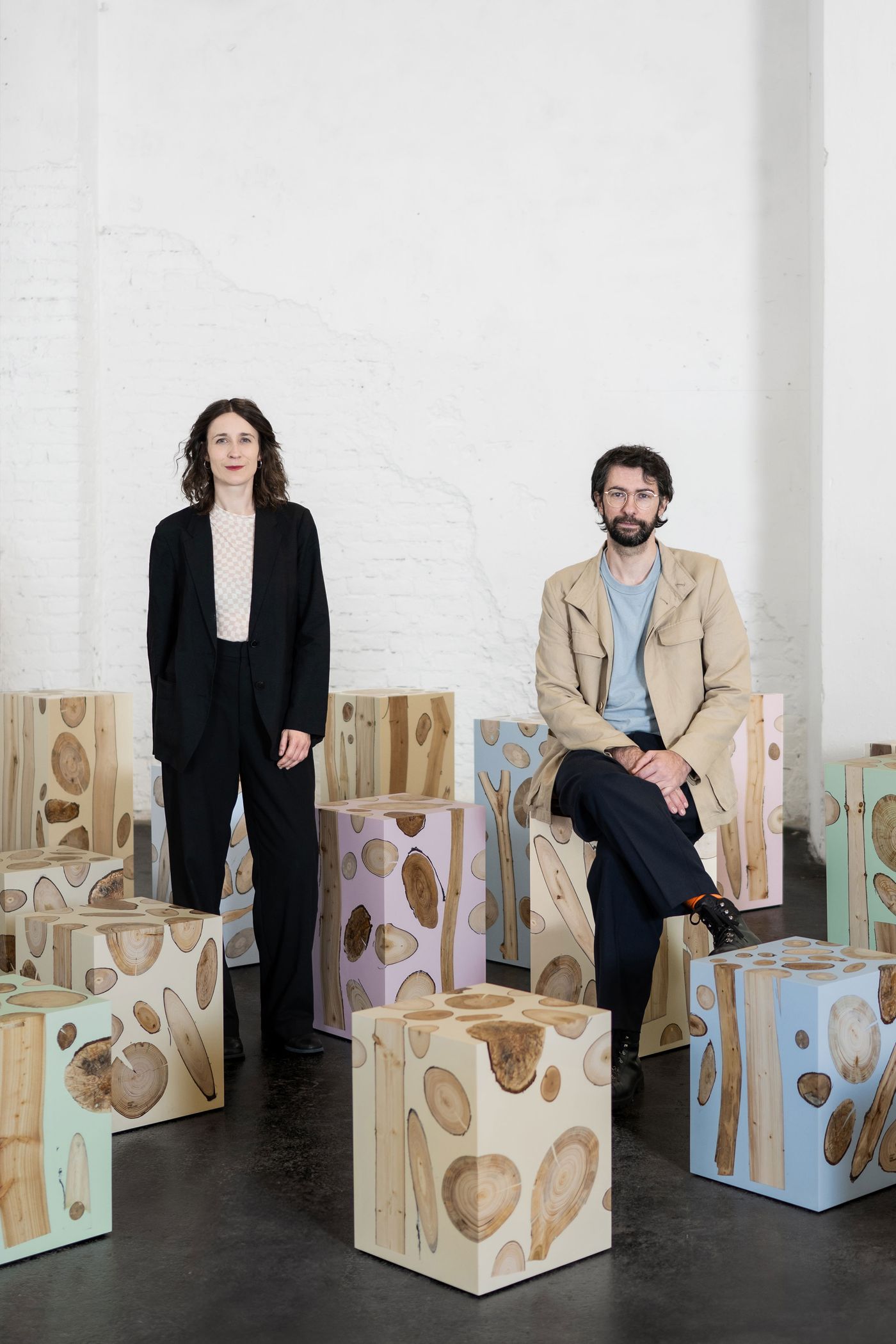 A man and a woman stand amidst an arrangement of cube-shaped stools. These stools are made from wood slices encased in clear resin, with some surfaces lightly colored in pastel shades of pink, blue, and green. The man is seated on one stool, while the woman stands beside another. The background is a white brick wall, suggesting an art gallery or studio space. The overall aesthetic is contemporary and artistic, highlighting natural materials in a unique application.