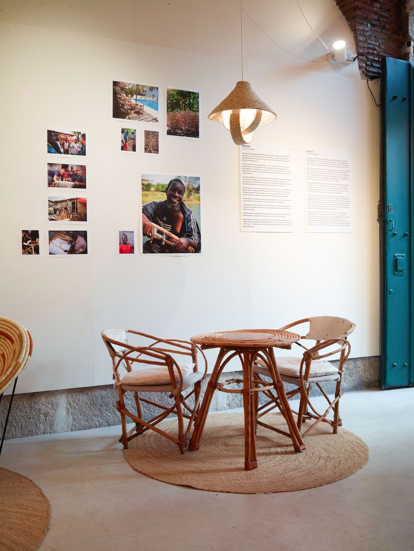 A corner of the "Kukua" exhibition at Prime Matter Gallery features a cozy natural rattan bistro set with a circular table and two chairs. Above, a sculptural woven pendant light illuminates a wall adorned with documentary-style photographs depicting people and craftsmanship. Adjacent information panels provide context, all set against ancient stone walls, merging contemporary design with cultural narrative.