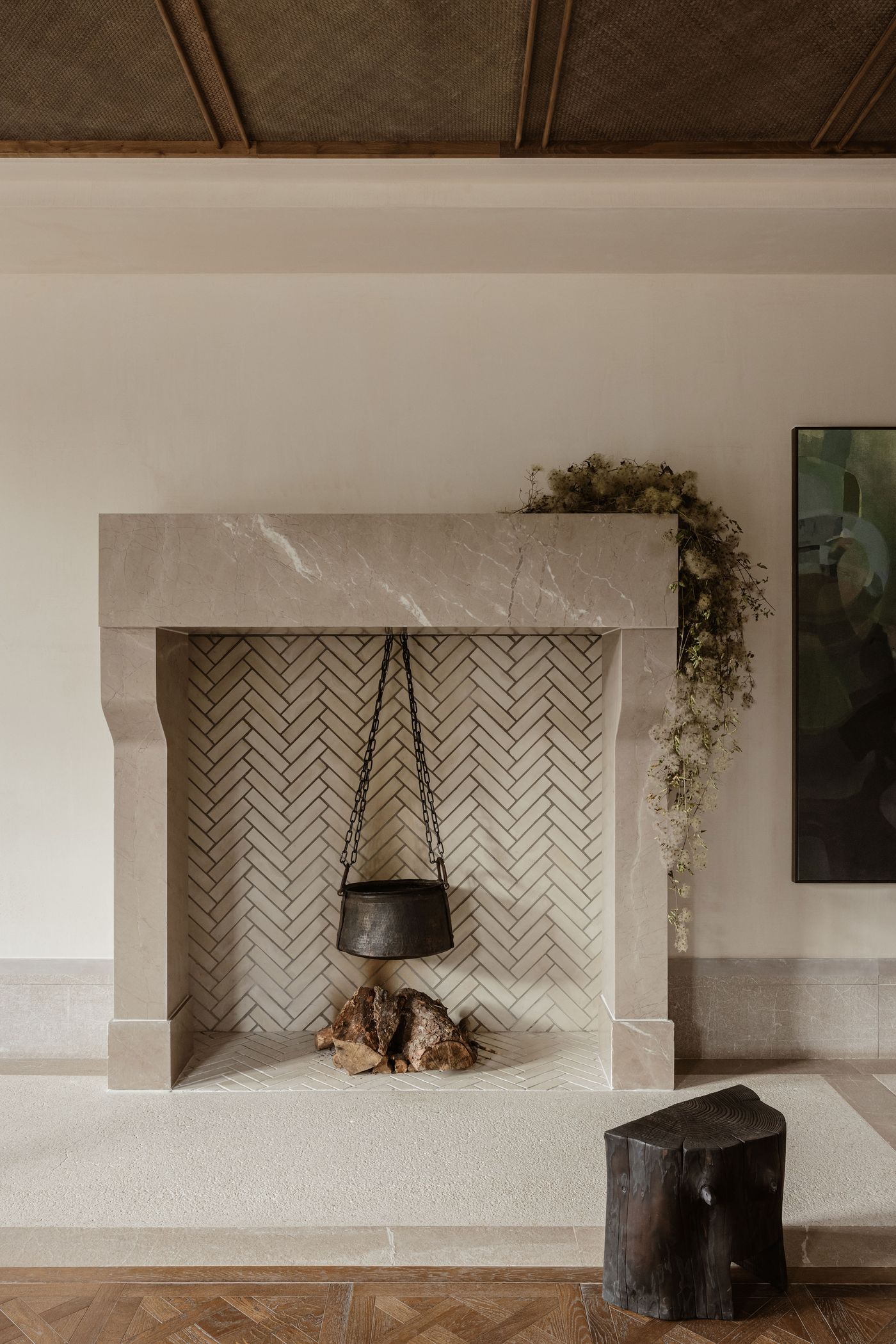 A robust, pale marble fireplace with a herringbone-patterned tile interior serves as a focal point, featuring a rustic black cauldron suspended over decorative firewood. An organic cascade of dried white flowers drapes from one side, adding a delicate, biophilic touch and blending modern elegance with traditional elements. A robust, pale marble fireplace with a herringbone-patterned tile interior serves as a focal point, featuring a rustic black cauldron suspended over decorative firewood. An organic cascade of dried white flowers drapes from one side, adding a delicate, biophilic touch and blending modern elegance with traditional elements.