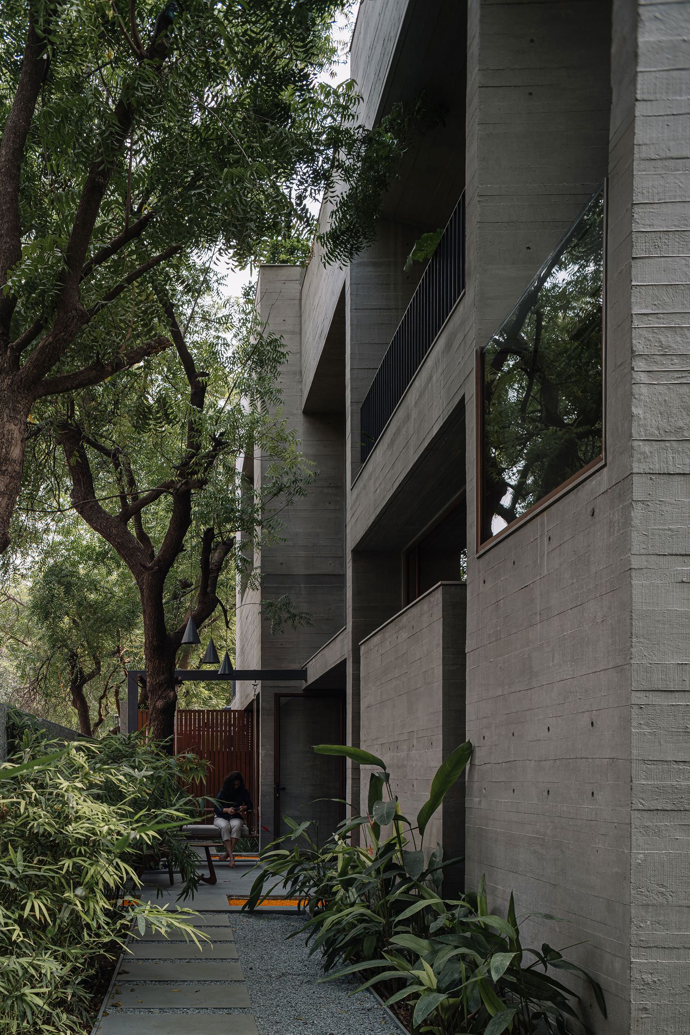 The exterior of MS House features multi-layered concrete facades with exposed formwork textures. Lush green trees and cascading vines soften the brutalist architecture, creating a natural canopy. A small outdoor seating area with people relaxing is visible on the ground level, demonstrating the seamless integration of indoor and outdoor living spaces.