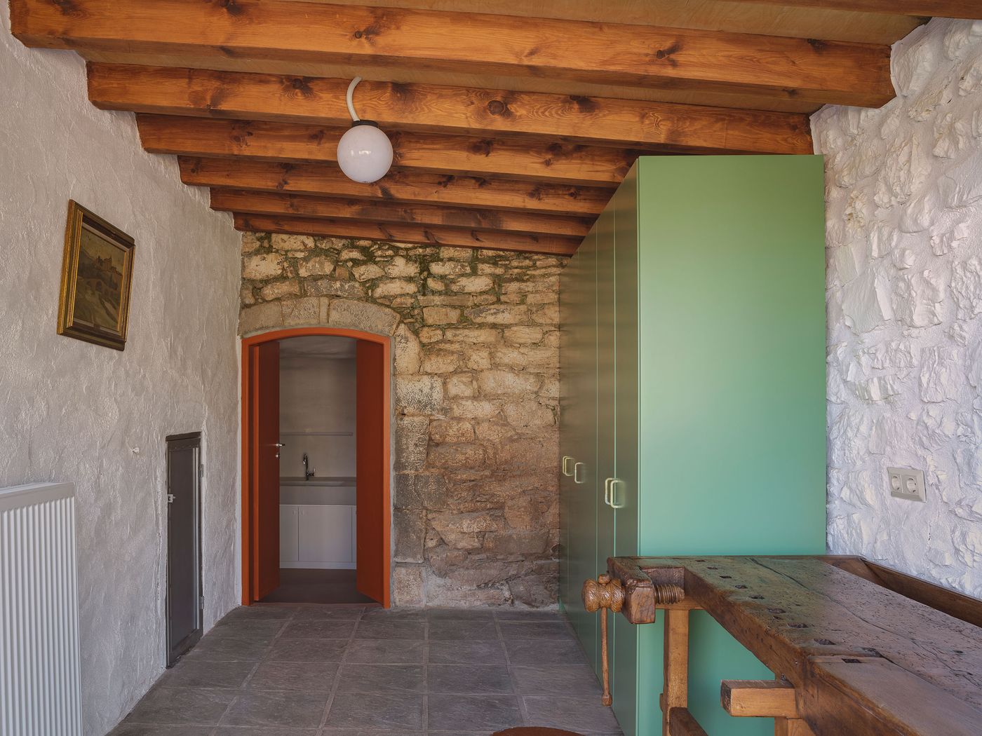Sage-green built-in storage creates functional room dividers beneath the traditional timber ceiling, where exposed stone walls display authentic Mediterranean texture and patina. Traditional wooden tools and furniture add rustic authenticity while contemporary cabinetry provides modern storage solutions within the preserved architectural shell.