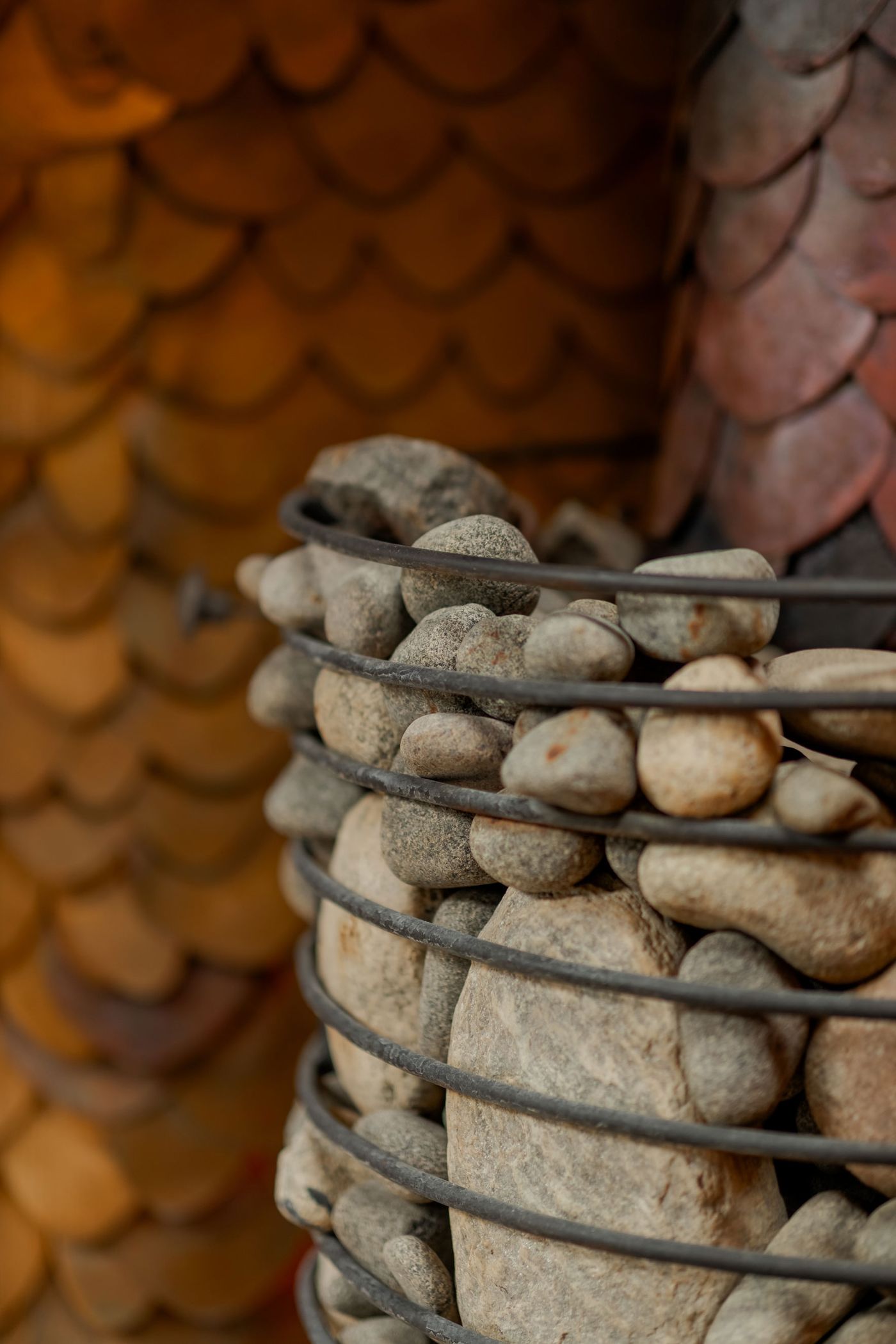 A close-up of a wire basket filled with smooth stones, set against a backdrop of textured, scale-like patterns in warm tones.