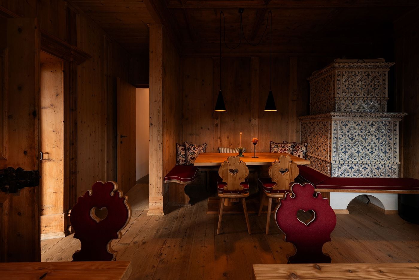 In a pine-panelled dining area at Castel Badia, Tyrolean chairs with heart-pierced backs, some upholstered in red felt, gather around a corner banquette. The tiled stove rises on a whitewashed arched base, its blue foliate tiles glowing beside a single candle, while an iron lock and doorways lead deeper into the castle. In a pine-panelled dining area at Castel Badia, Tyrolean chairs with heart-pierced backs, some upholstered in red felt, gather around a corner banquette. The tiled stove rises on a whitewashed arched base, its blue foliate tiles glowing beside a single candle, while an iron lock and doorways lead deeper into the castle.