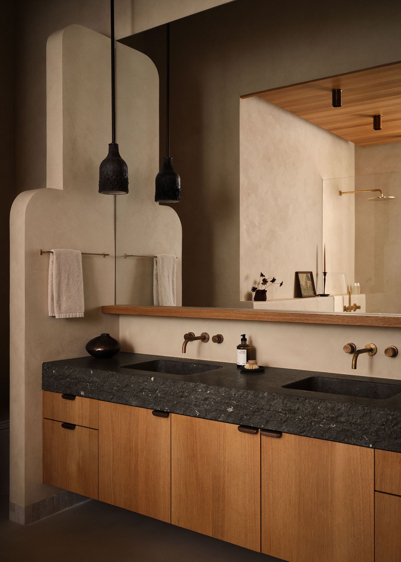Minimalist bathroom featuring dual sinks, wooden cabinetry, black pendant lights, and earthy tones, with stylish decor accents.