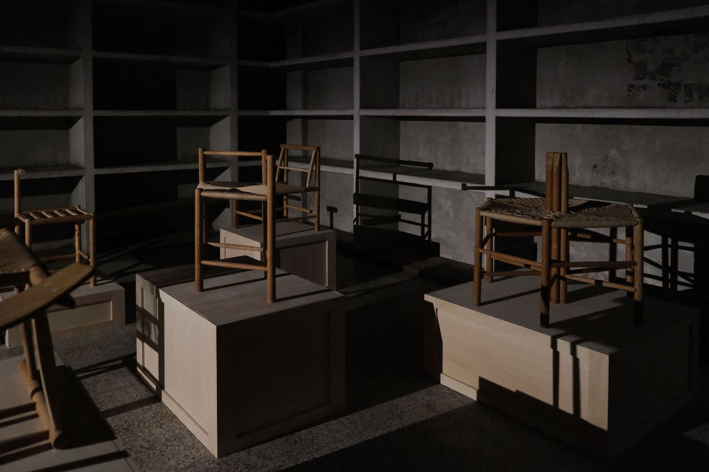 A dimly lit room with wooden chairs displayed on pedestals, surrounded by shadowy shelves and a concrete wall. A dimly lit room with wooden chairs displayed on pedestals, surrounded by shadowy shelves and a concrete wall.