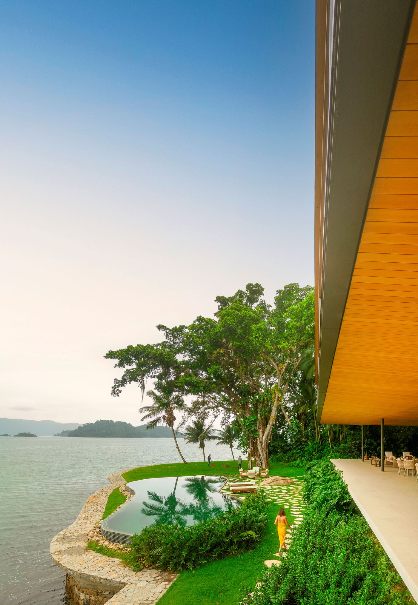 A person in a vibrant yellow dress walks along a stepping stone path, leading towards an infinity pool that merges with the serene ocean. The cantilevered wooden structure above provides shelter, while towering tropical trees frame the view, creating a tranquil and luxurious outdoor living space that embraces the coastal landscape.