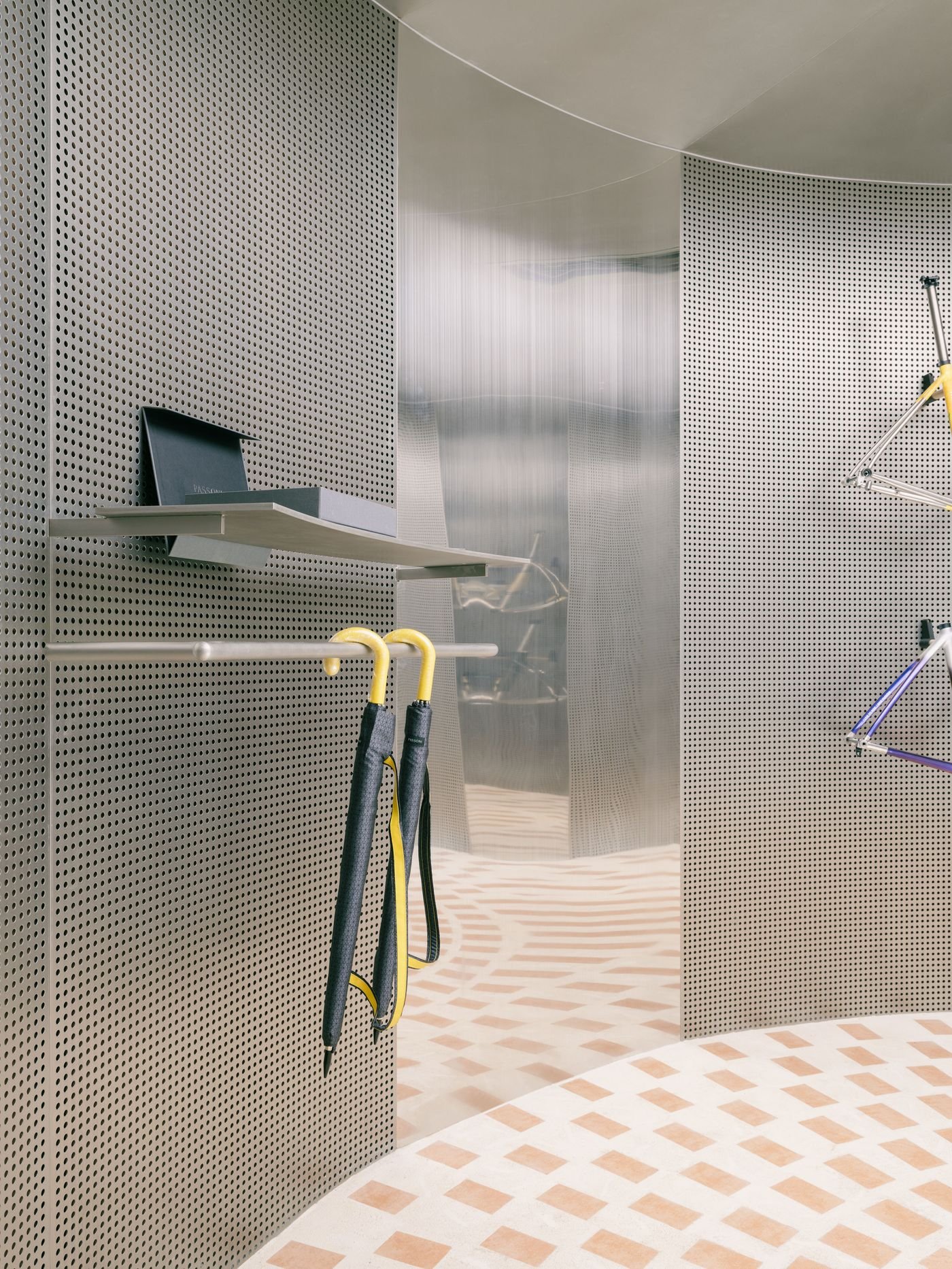 A close-up view reveals the intricate details and textures within AITASHOP, featuring a wall of light grey perforated metal that serves as a display surface with a minimalist shelf. Two umbrellas with vibrant yellow handles hang from a bar, introducing a pop of color against the neutral, industrial palette. The circular terracotta-tiled floor creates a soft, inviting pattern. A close-up view reveals the intricate details and textures within AITASHOP, featuring a wall of light grey perforated metal that serves as a display surface with a minimalist shelf. Two umbrellas with vibrant yellow handles hang from a bar, introducing a pop of color against the neutral, industrial palette. The circular terracotta-tiled floor creates a soft, inviting pattern.